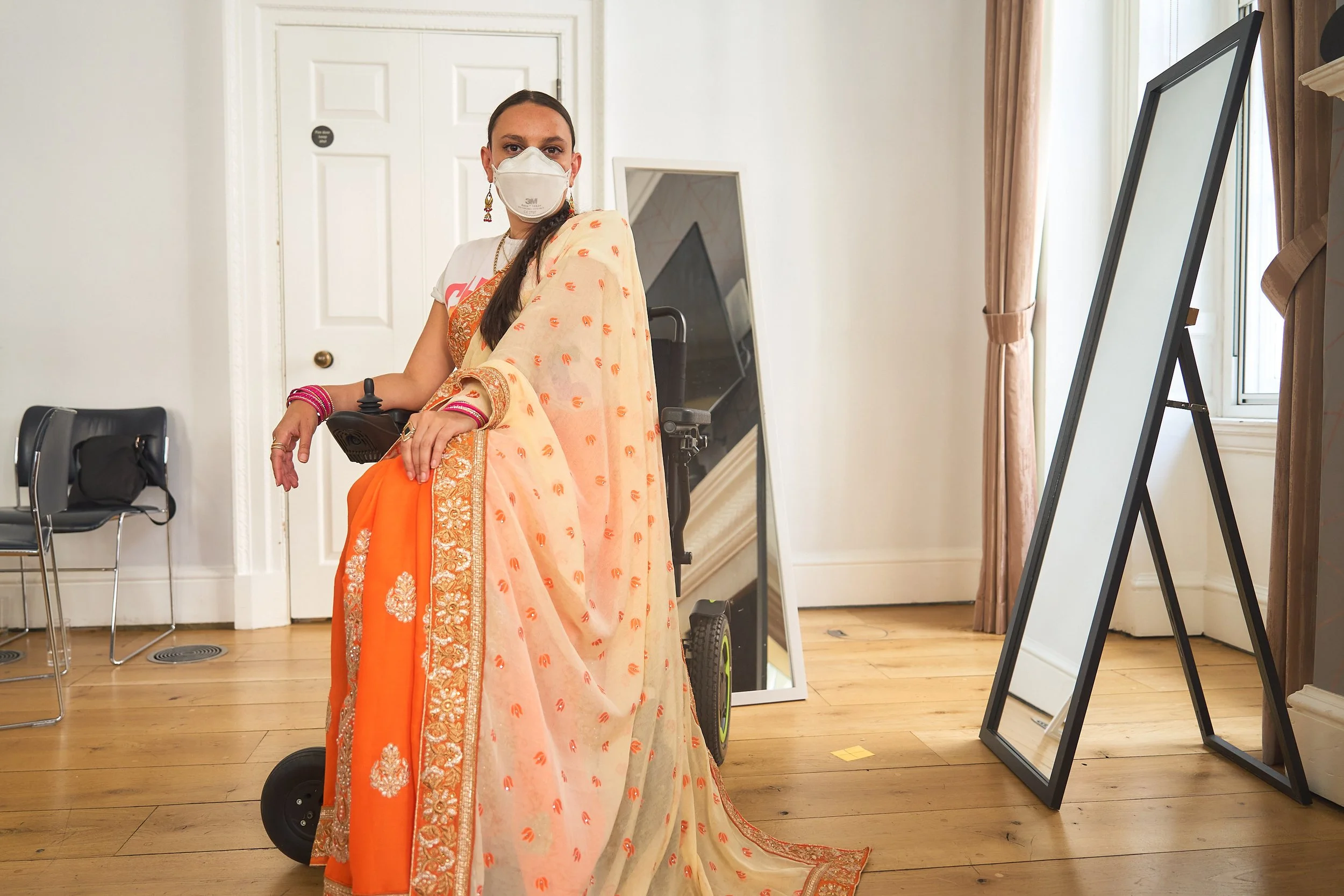 A woman wearing an orange and gold patterned saree sits in a wheel chair, with two mirrors behind her