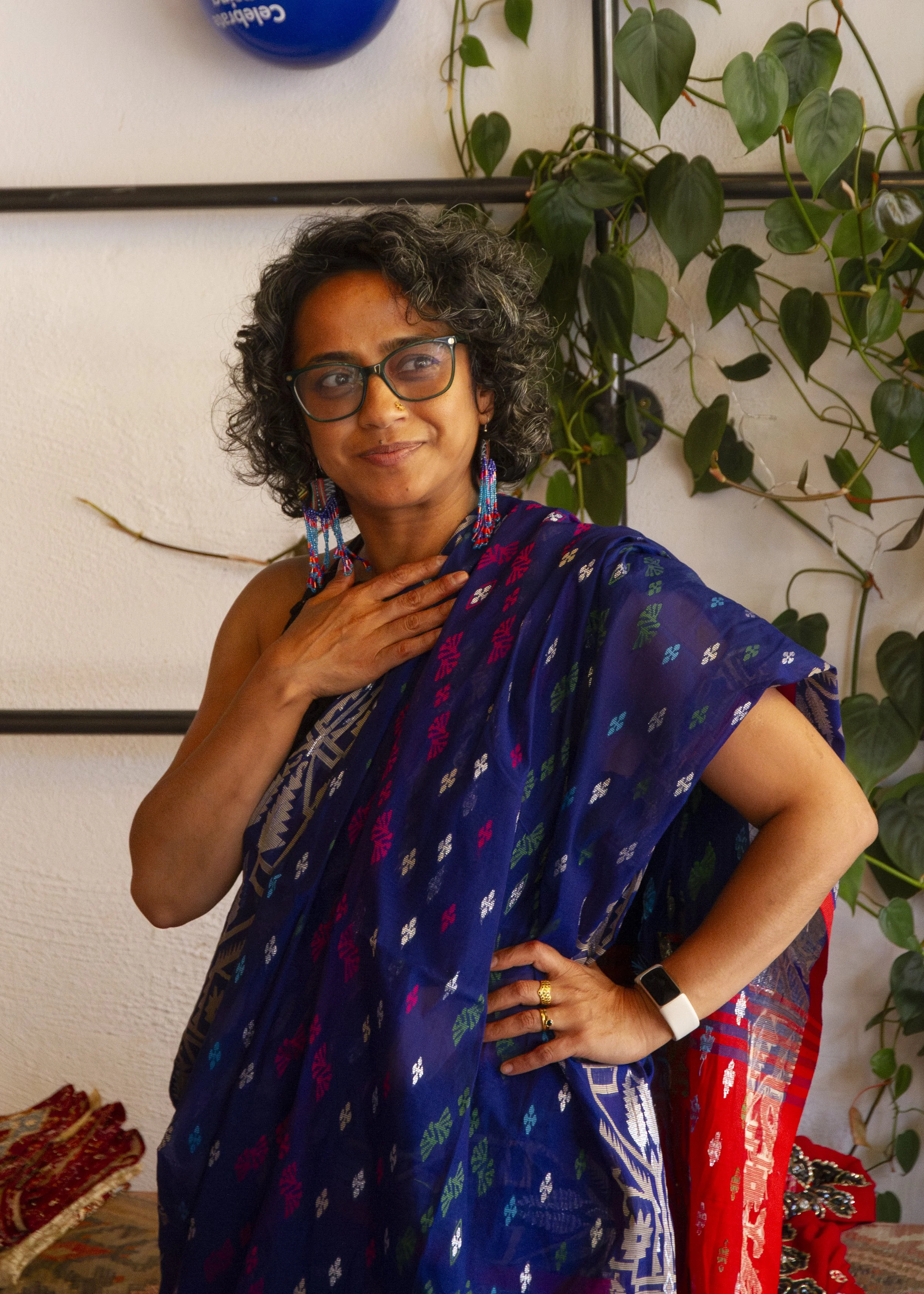A south Asian woman with curly hear wearing a blu saree stands straight with one hand on her chest and the other hand on her hip
