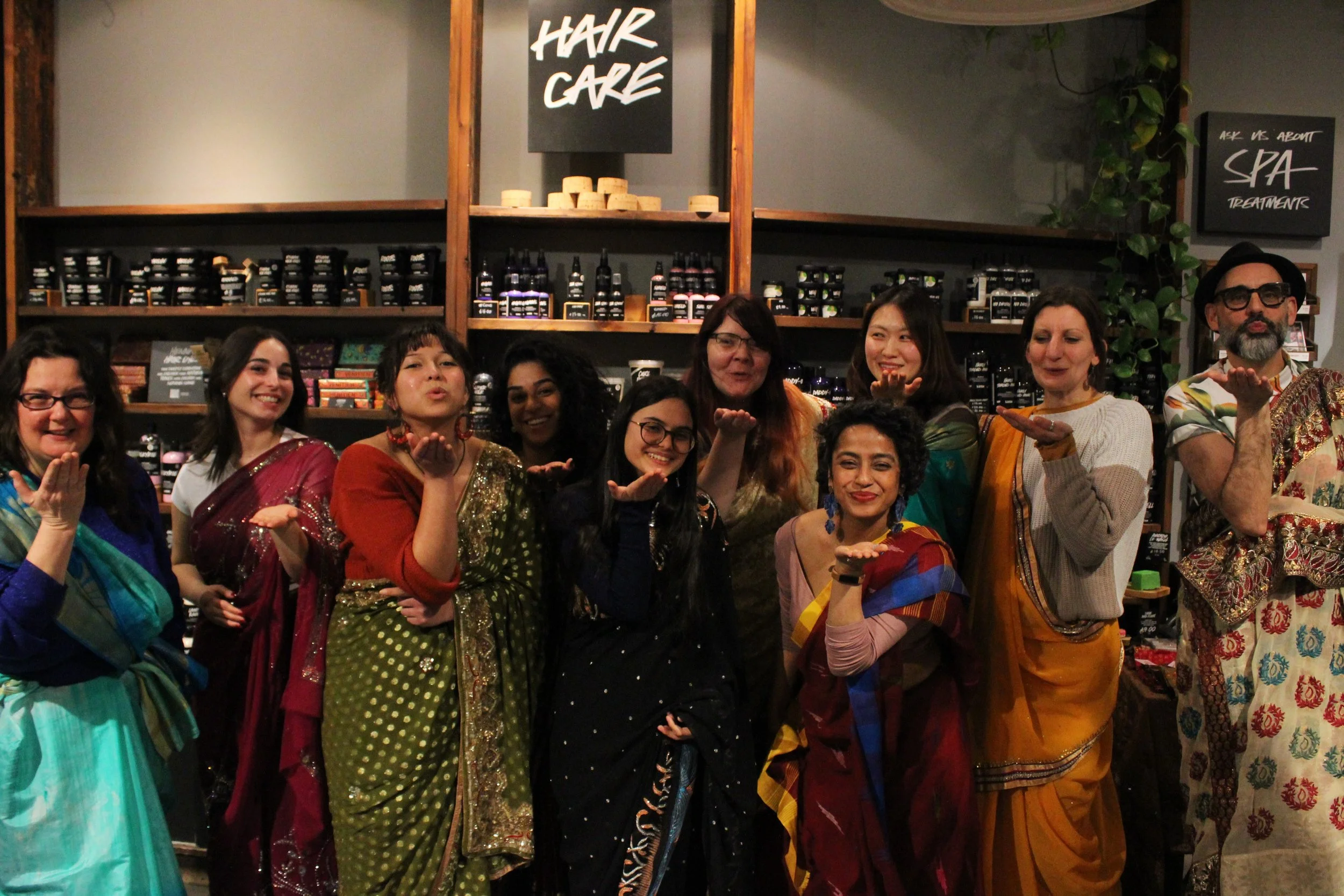 A group of people draped in sarees stand in a line and blow a kiss toward the camera