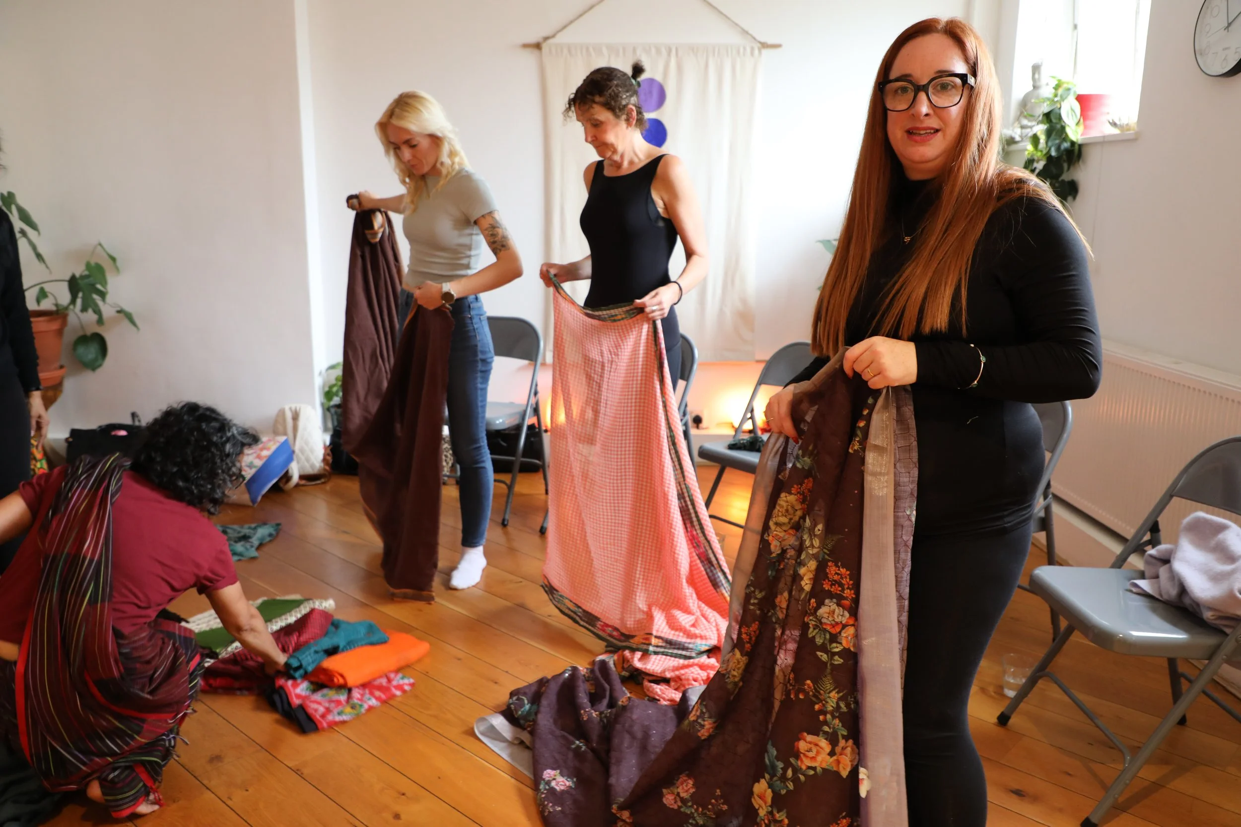 Three women hold lengths of saree fabric in their hands to wrap it around themselves. Another women with curly hair squats on the floor to pick up a saree from the floor
