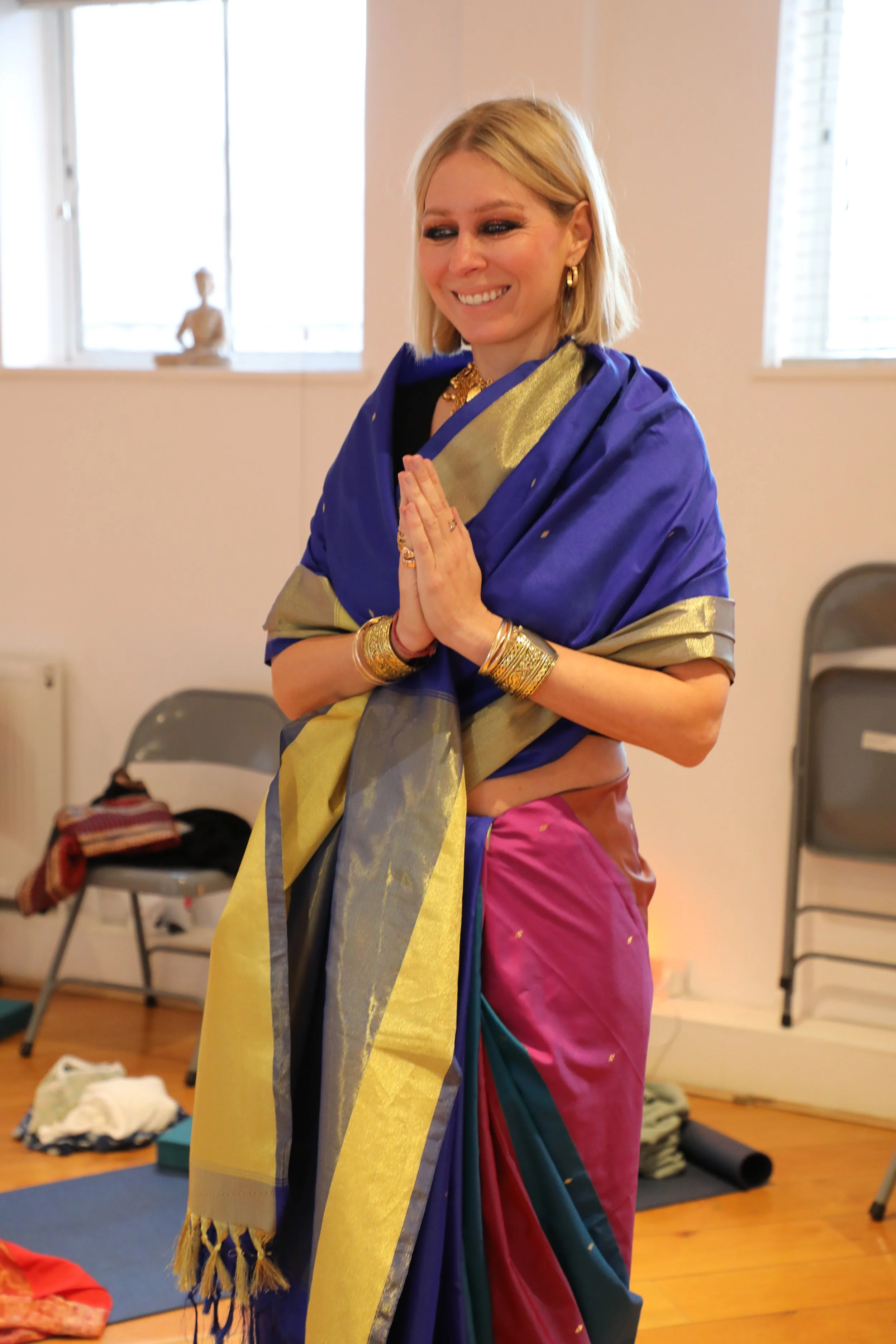 A front view of a woman wearing a colourful saree with her hands placed together in a prayer position, smiling and not looking at the camera