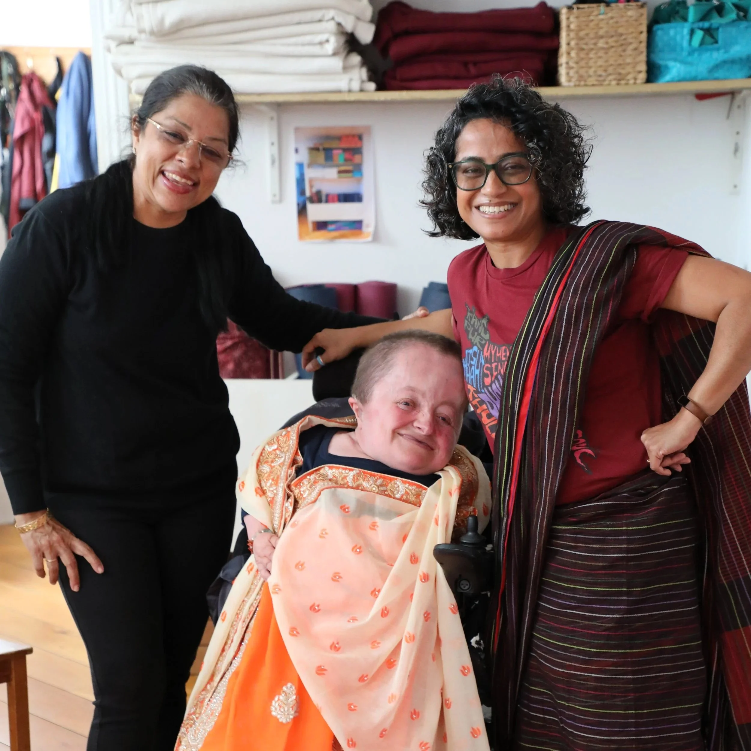 Two women stand on either side of a woman sitting in a power wheelchair, all smiling at the camera