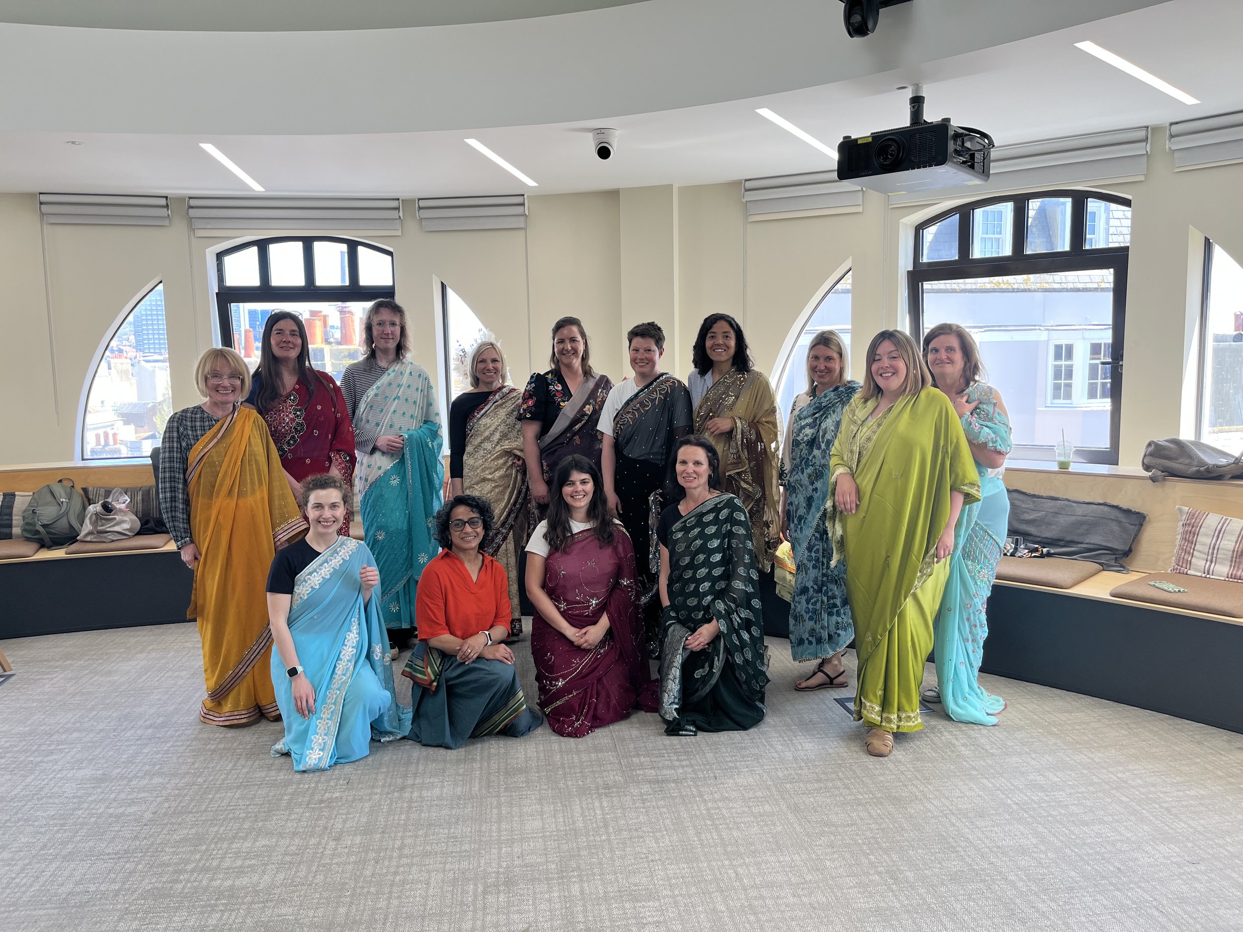 10 women wearing colourful sarees stand together with four women kneeling in front of them, all looking toward the camera and smiling