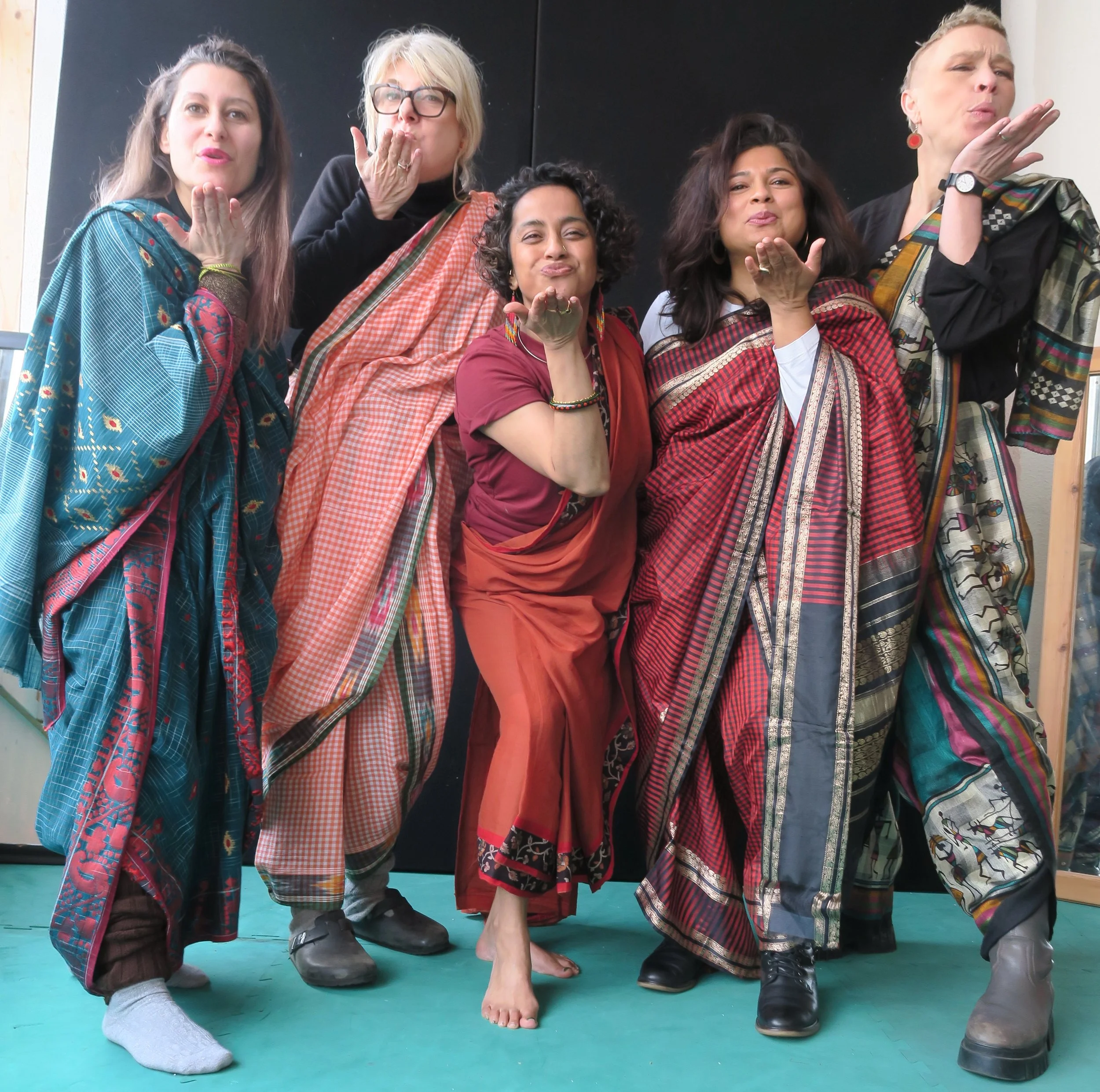 Five women wearing colourful sarees stand together blowing a kiss to the camera