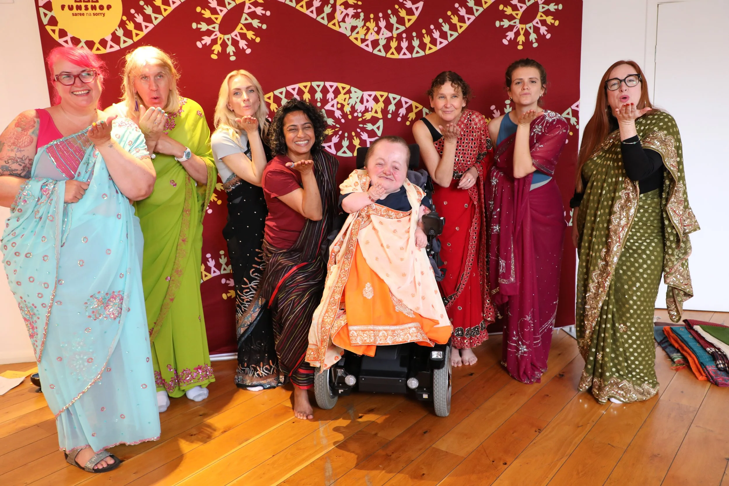 Eight women wearing sarees all blow a kiss at the camera