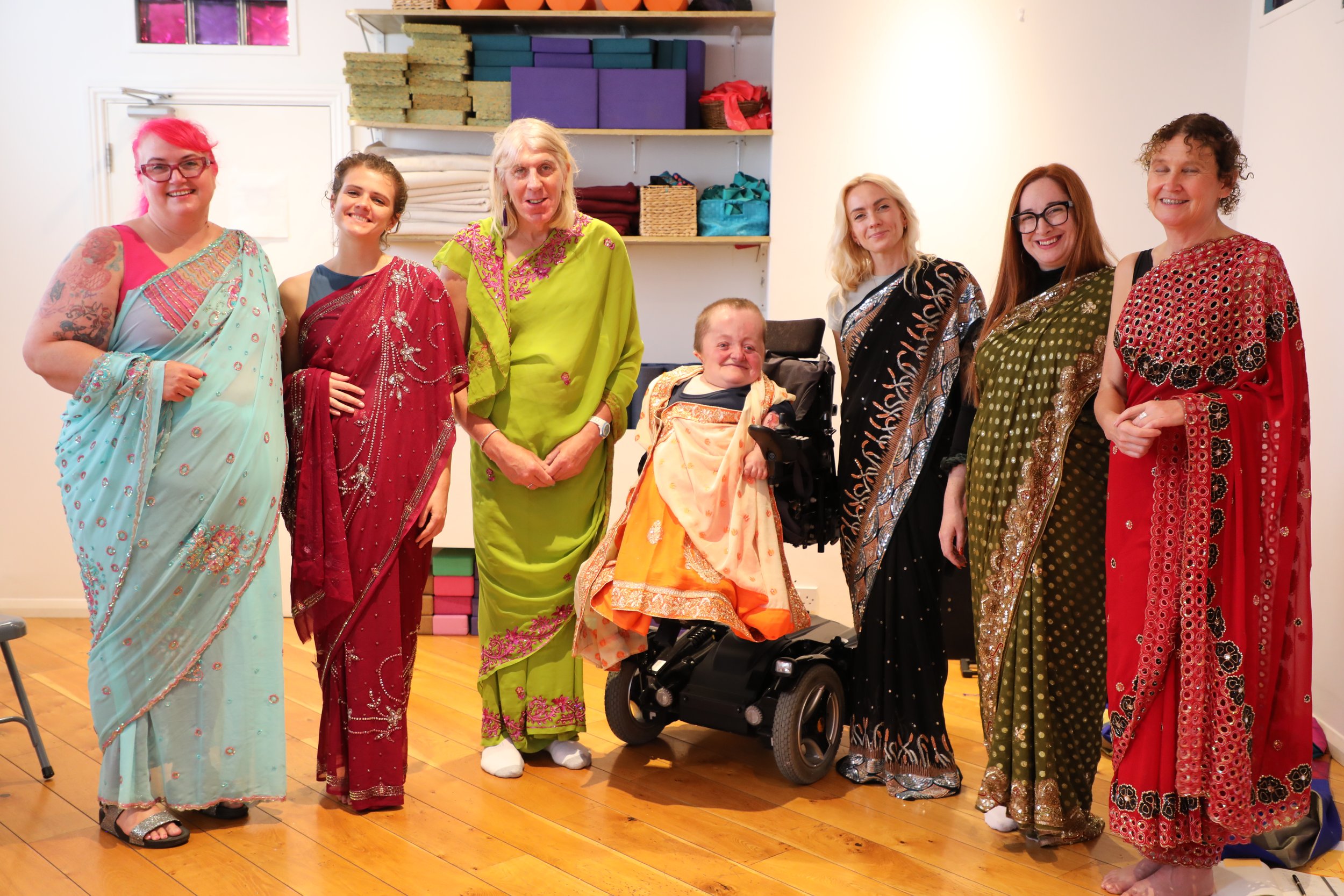 Seven people wearing colourful sarees arranged in a line formation smile toward the camera