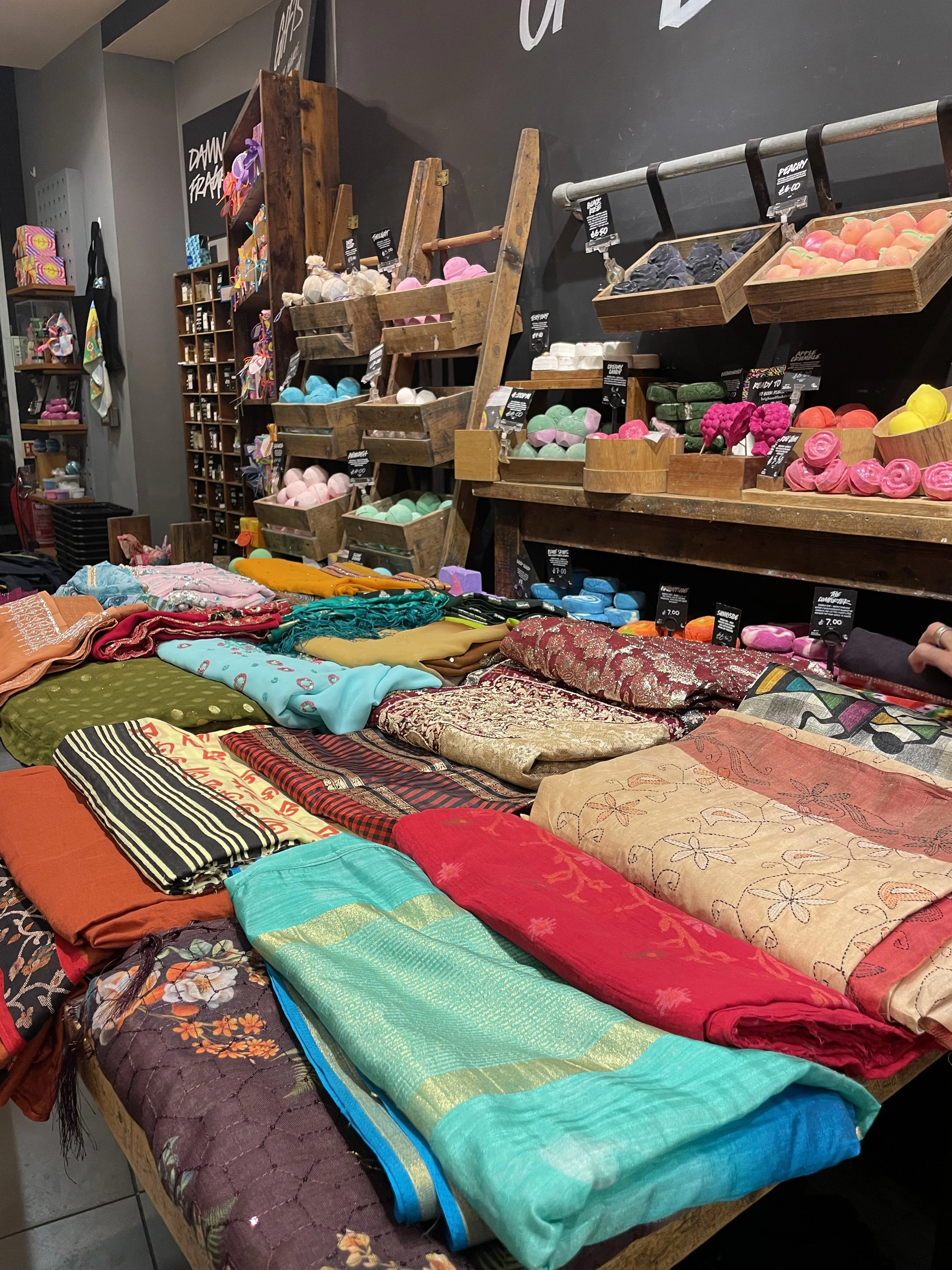 A table full of colourful sarees in front of a wall lined with colourful bath bombs