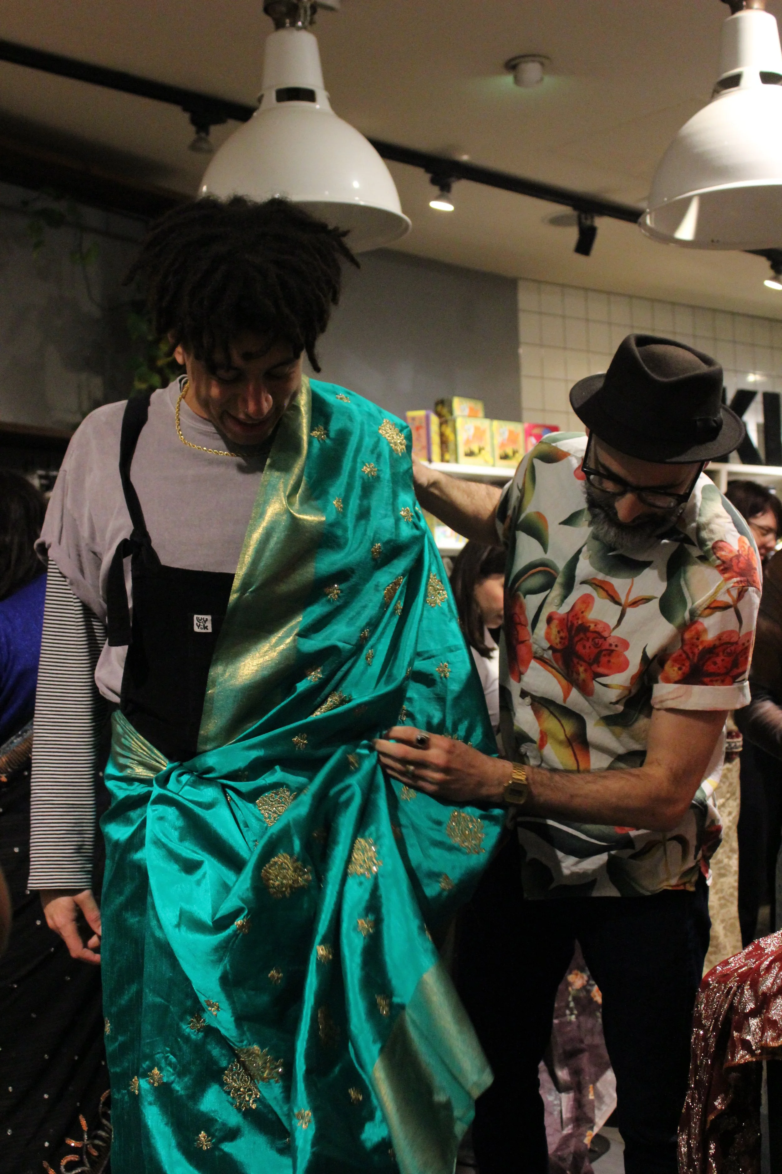 Two men looking away from the camera, one helps the other drape in a green saree