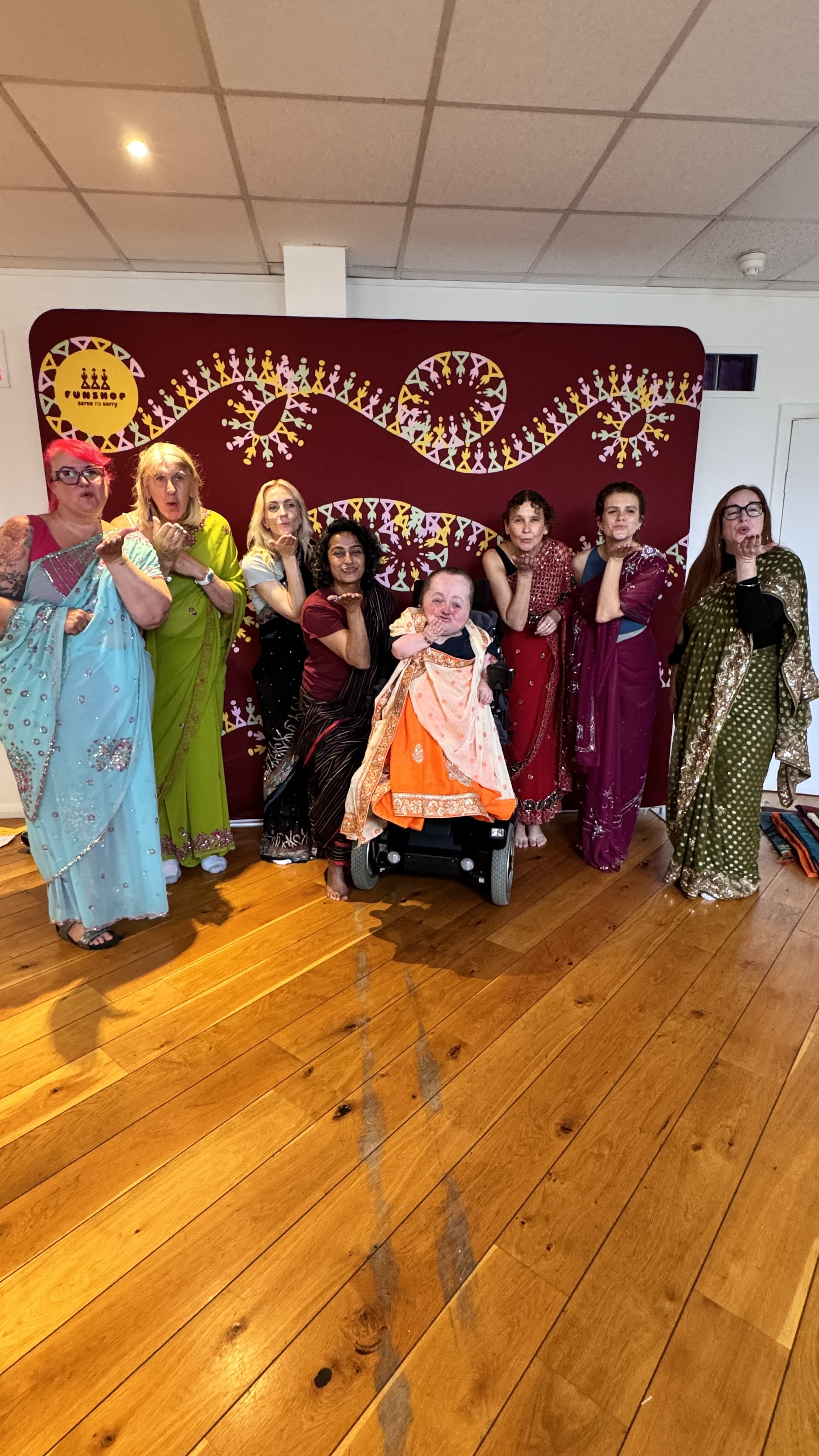 eight people wearing brightly coloured sarees stand in front of a colourful backdrop and blow a kiss at the camera