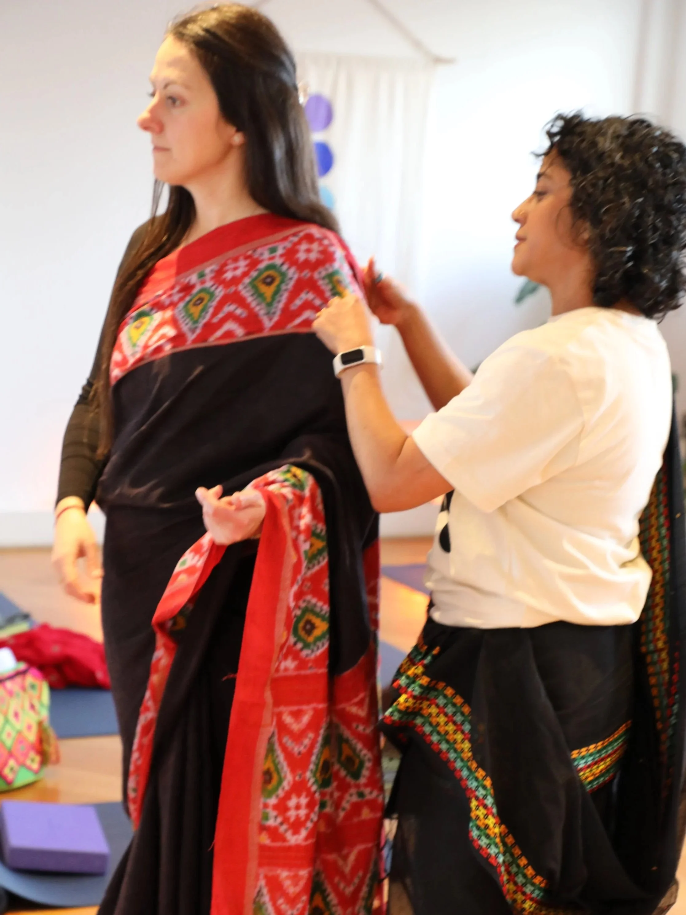 A woman wearing a white t shirt and a black saree helps another woman wearing a black saree to drape it over her shoulder