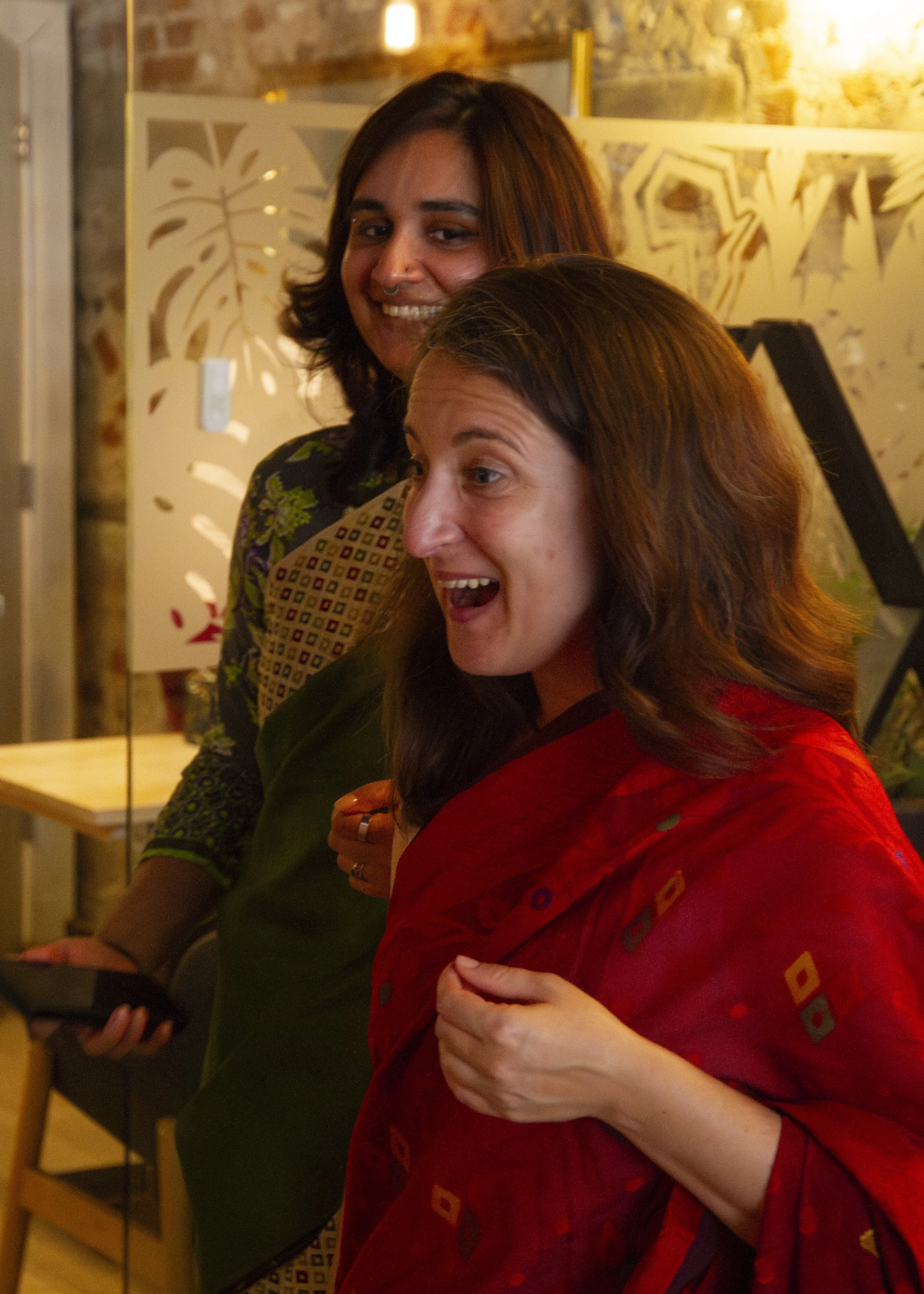 A white British woman with dark hair wearing a saree looks excited and a South Asian woman smiles from behind her