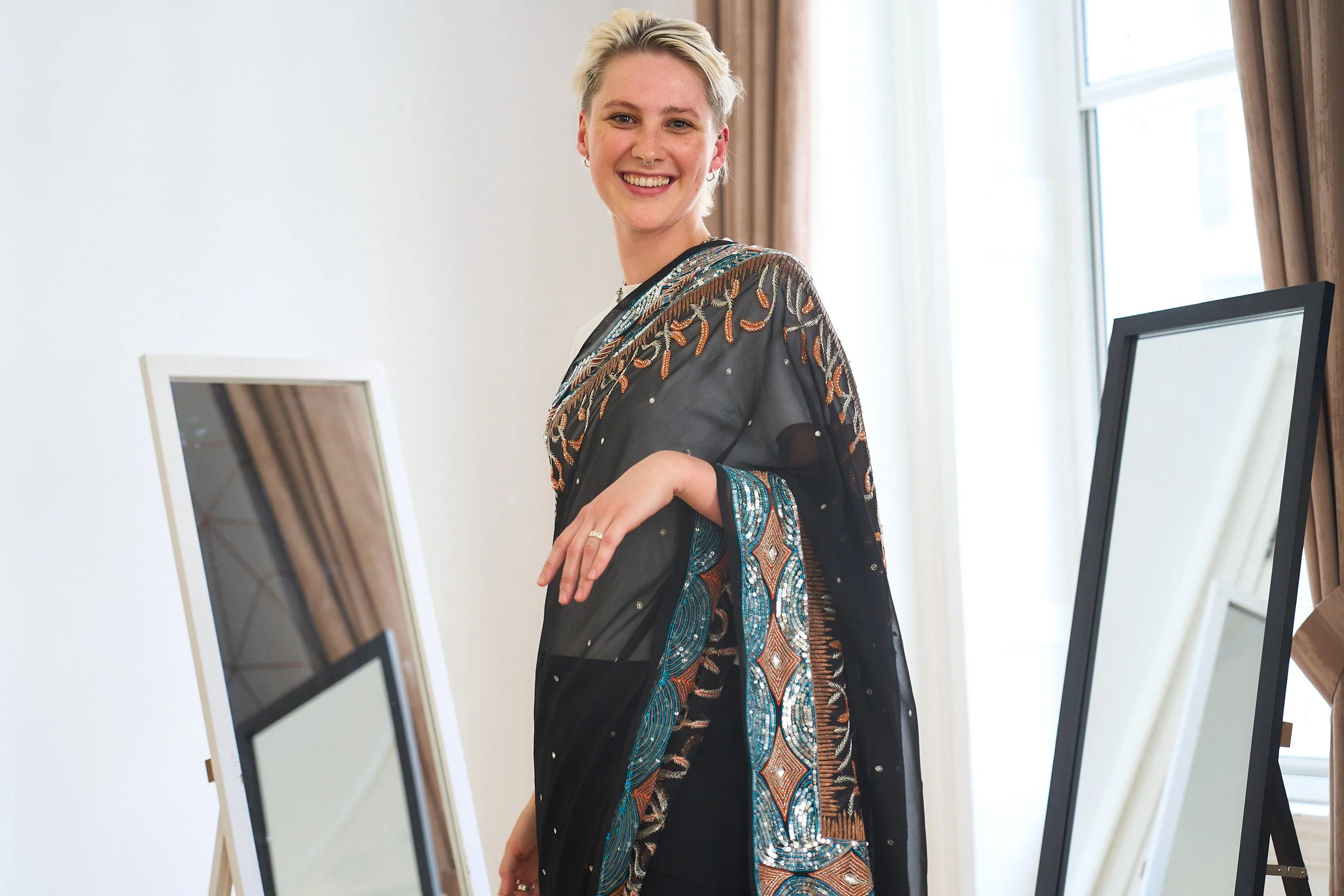 A white British woman wearing a black embellished saree smiles at the camera, with two mirrors behind her