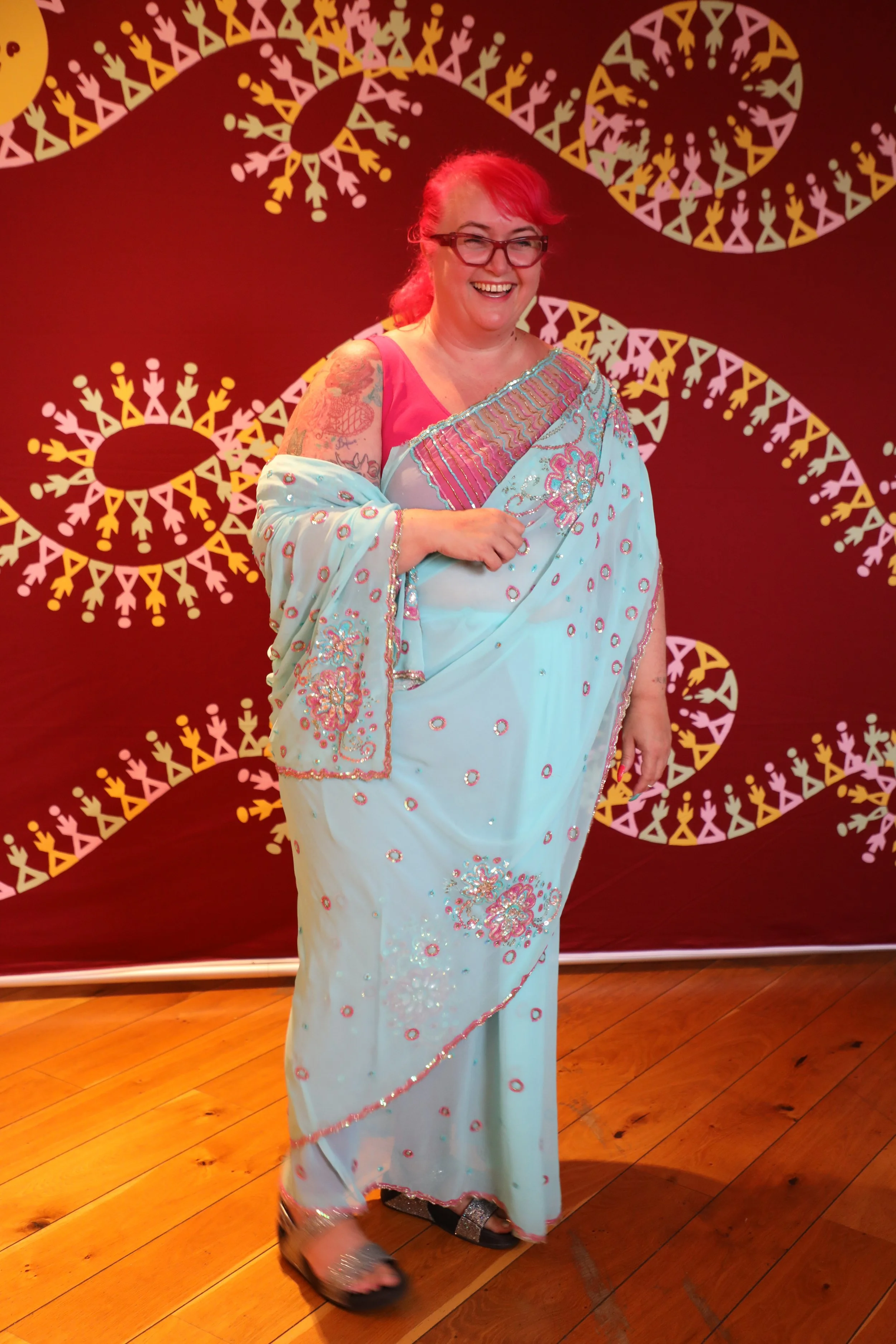 A white british woman wearing glasses and a blue patterned saree stands and smiles, looking away from the camera