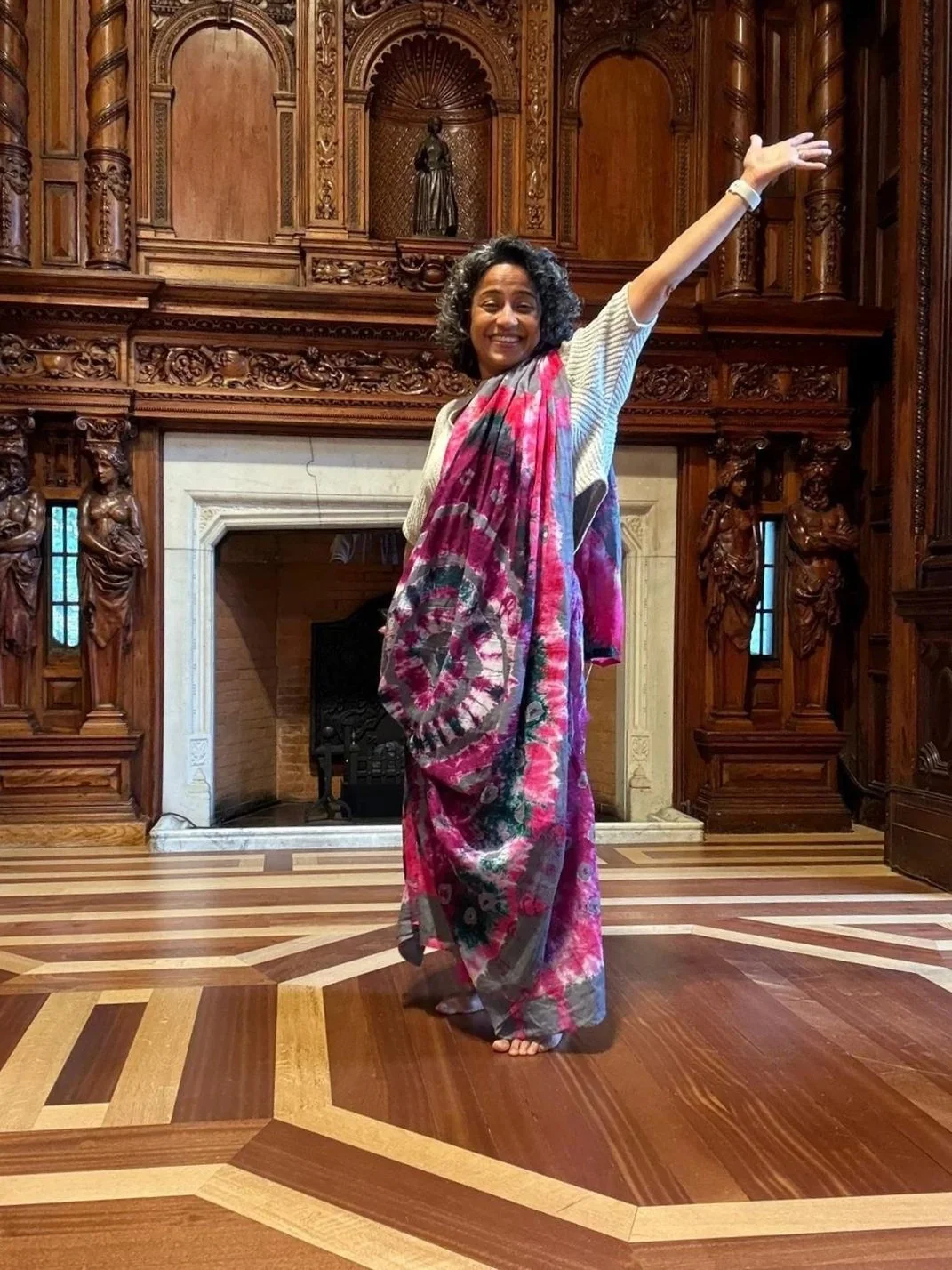 A British-Bangladeshi woman wearing a colourful pink saree stands in the foreground with one arm outstretched and smiles at the camera