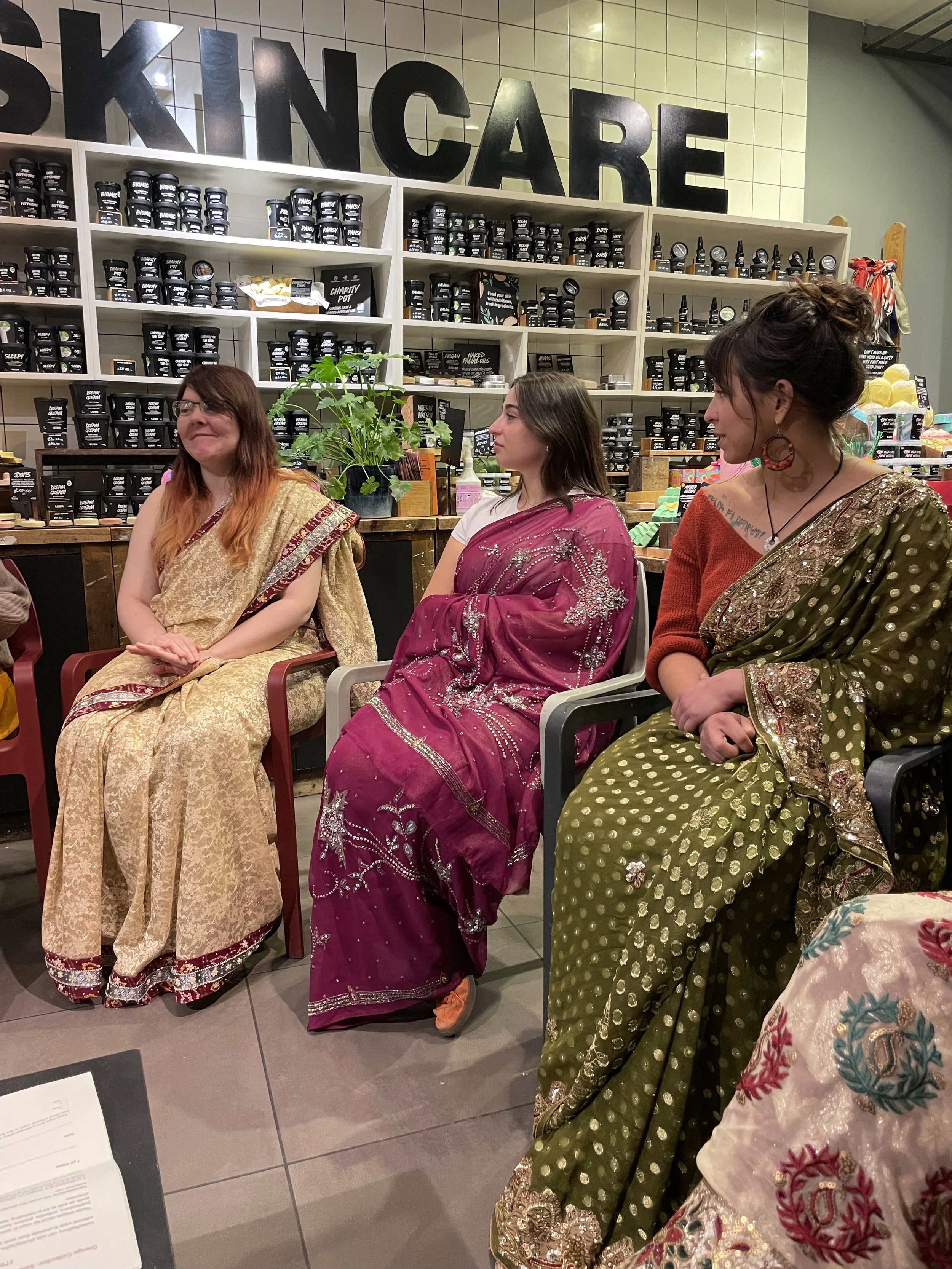 Three women wearing colourful sarees sit in chairs and talk