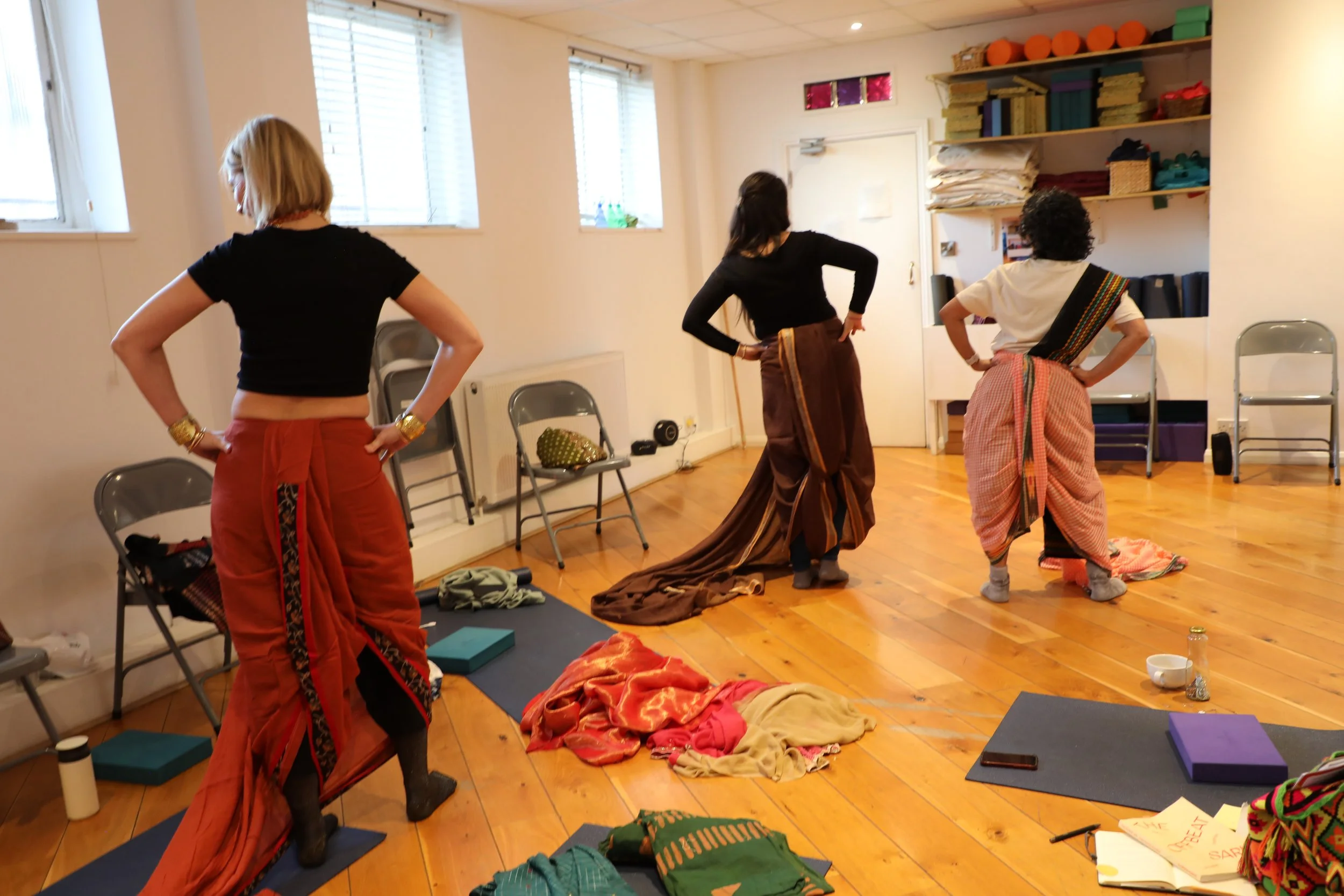 Three women wearing sarees in a trouser drape stand with their hands on their hips in a room full of sarees and yoga mats on the floor