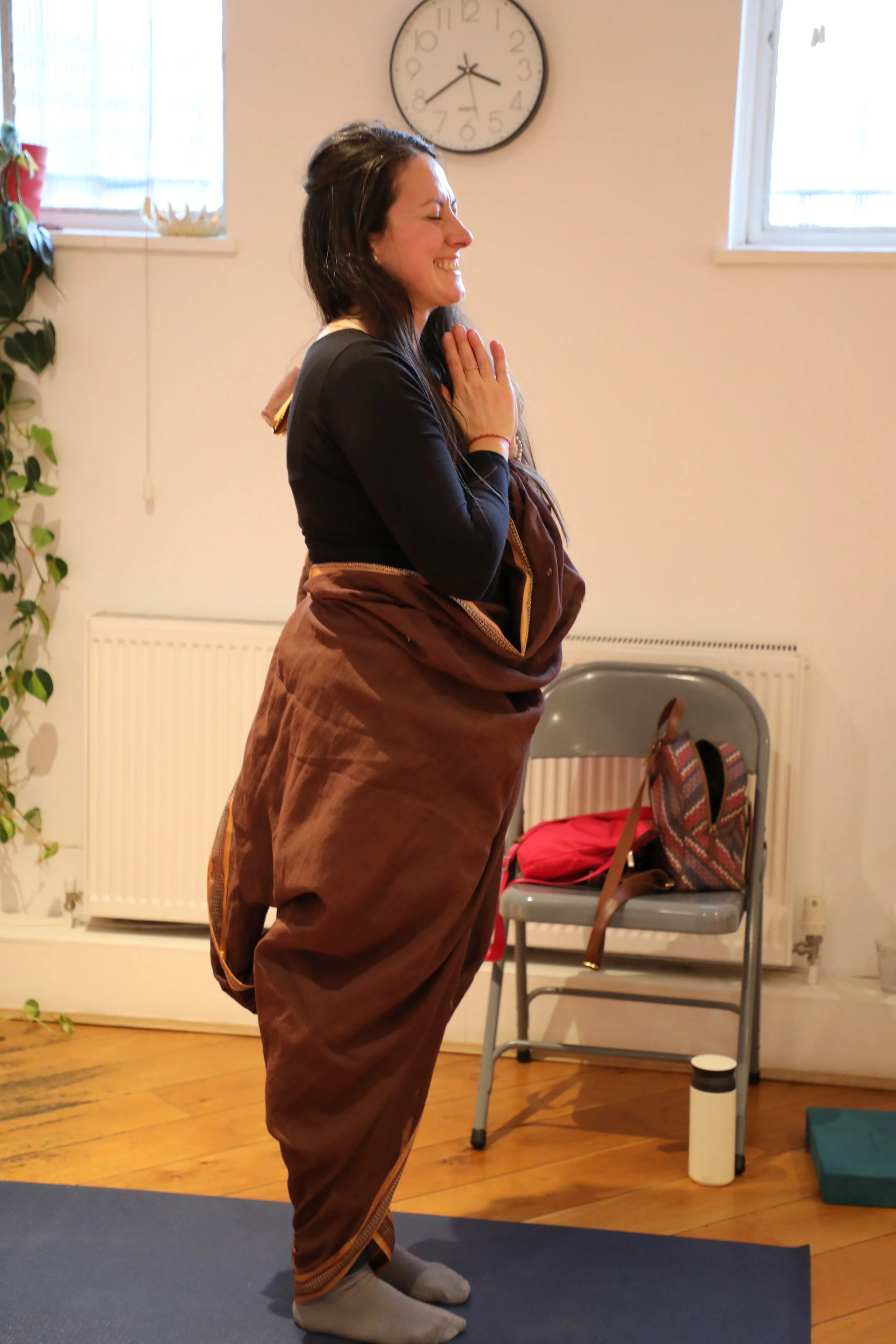 A woman wearing a brown saree draped in a trouser style stands with her hands together in a prayer position, smiling and looking away from the camera