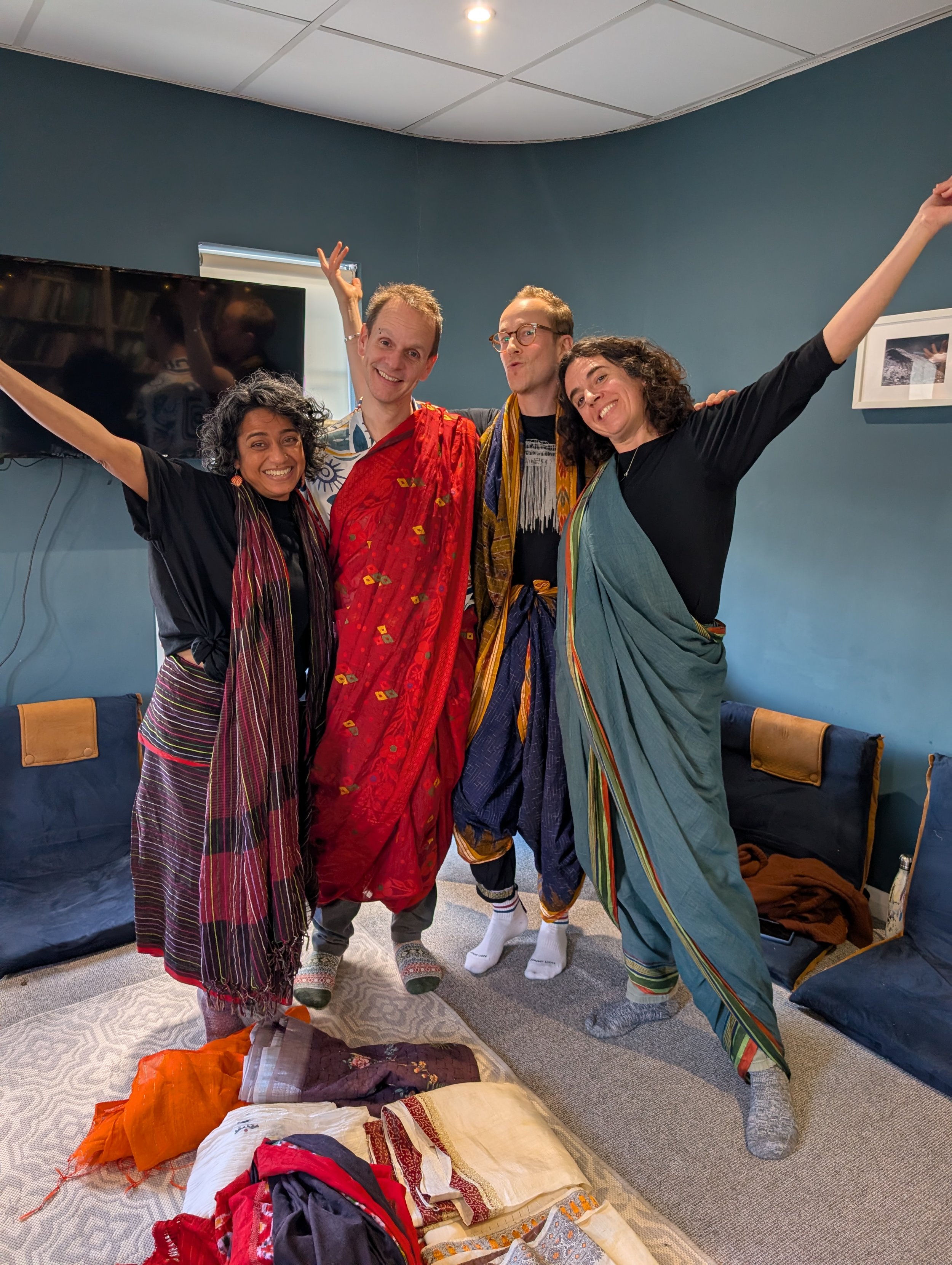4 people stand together with their arms outstretched, dressed in differently coloured sarees
