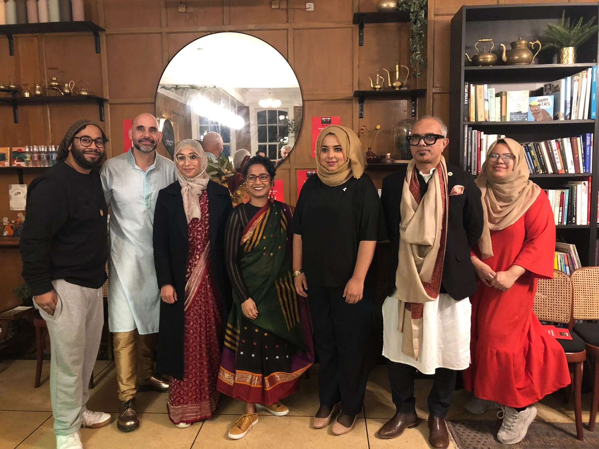A group of 6 people are standing grouped together smiling. There is a mix of genders and ethnicities and two of the women are wearing sarees. 