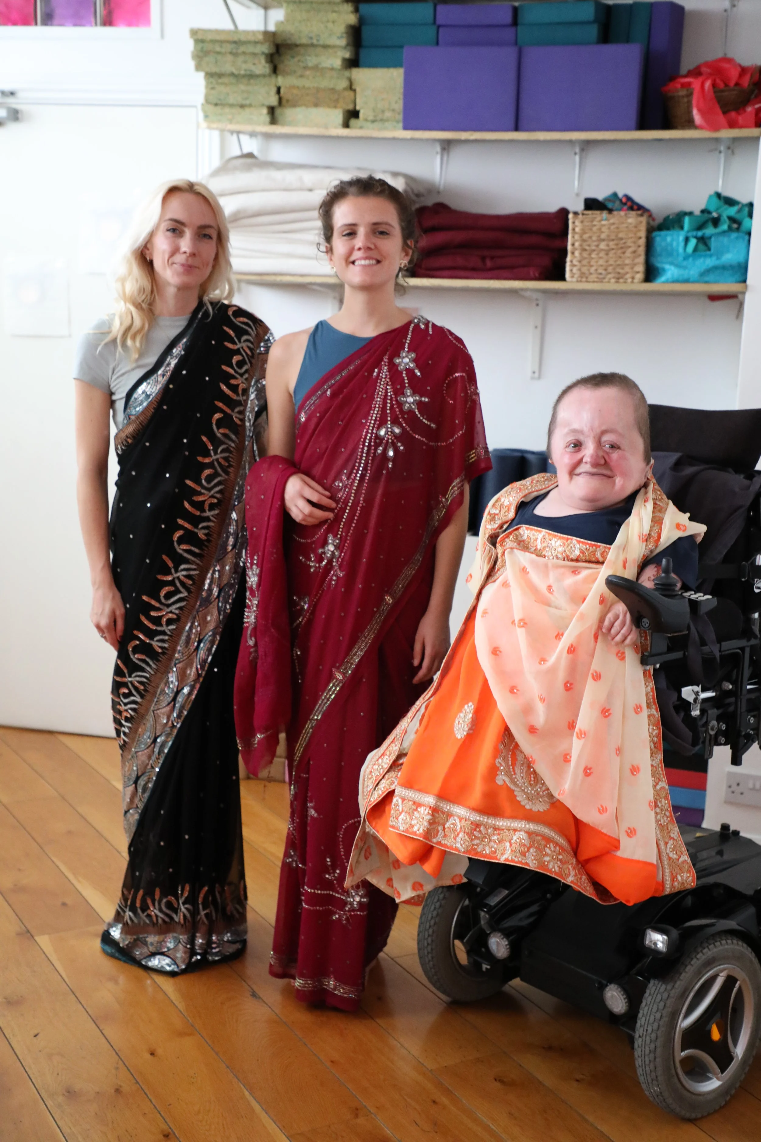 Two white British women wearing sarees smile and stand next to another white British person in a power wheelchair wearing an orange saree