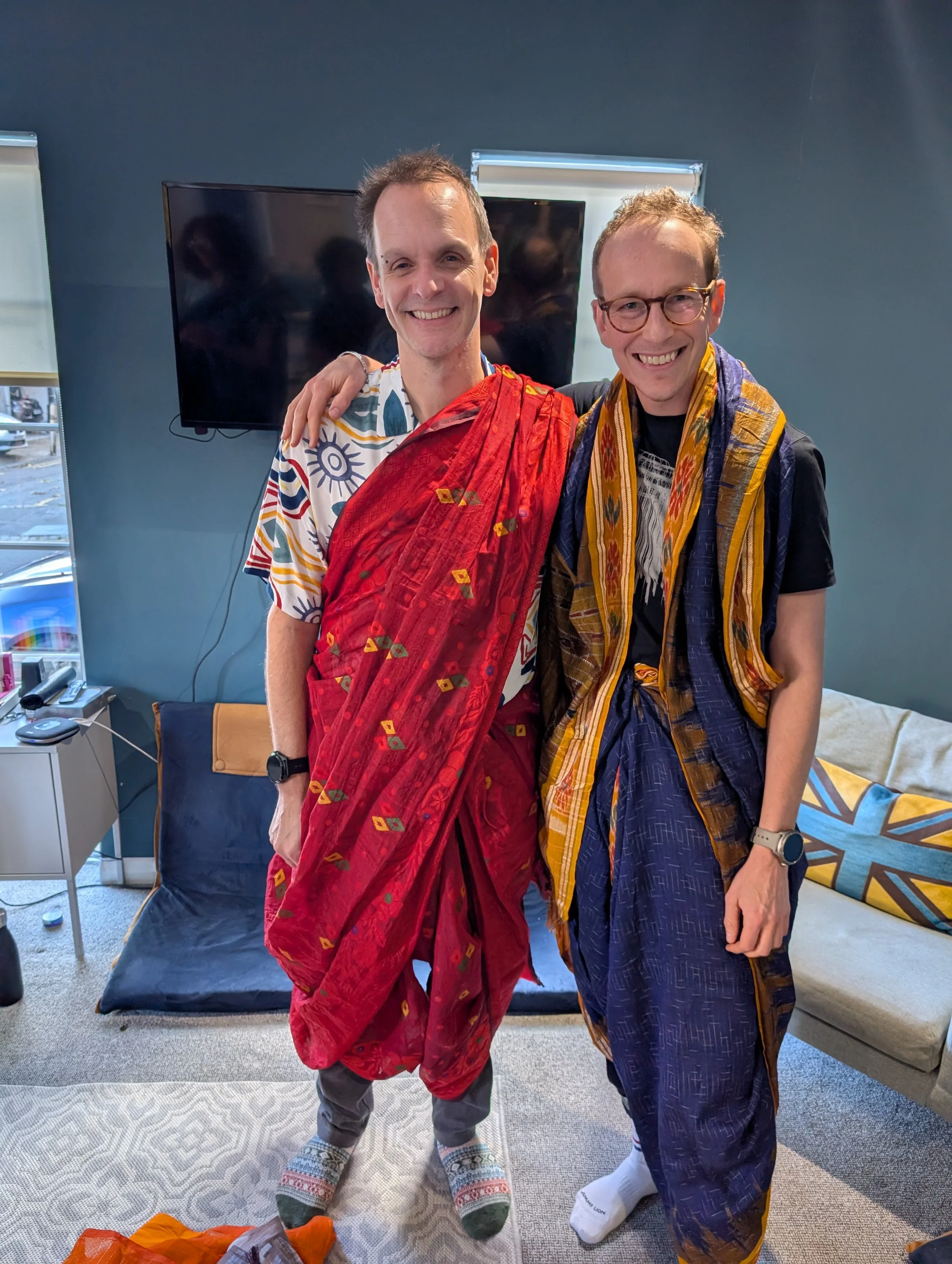 Two men wearing sarees in different drapes smile at the camera with their arms around each others shoulders