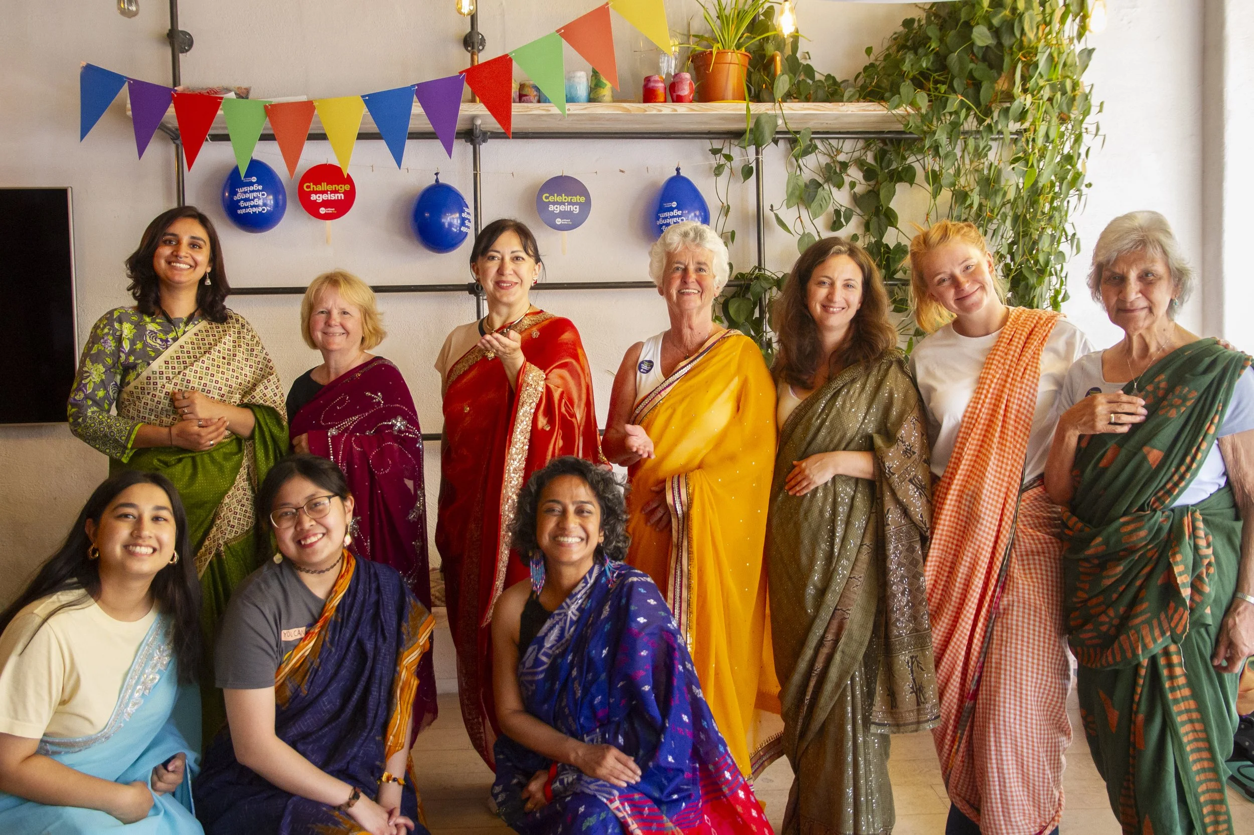 ten women of varying ages and races smile at the camera. All are dressed in sarees, three women crouch while seven stand straight.