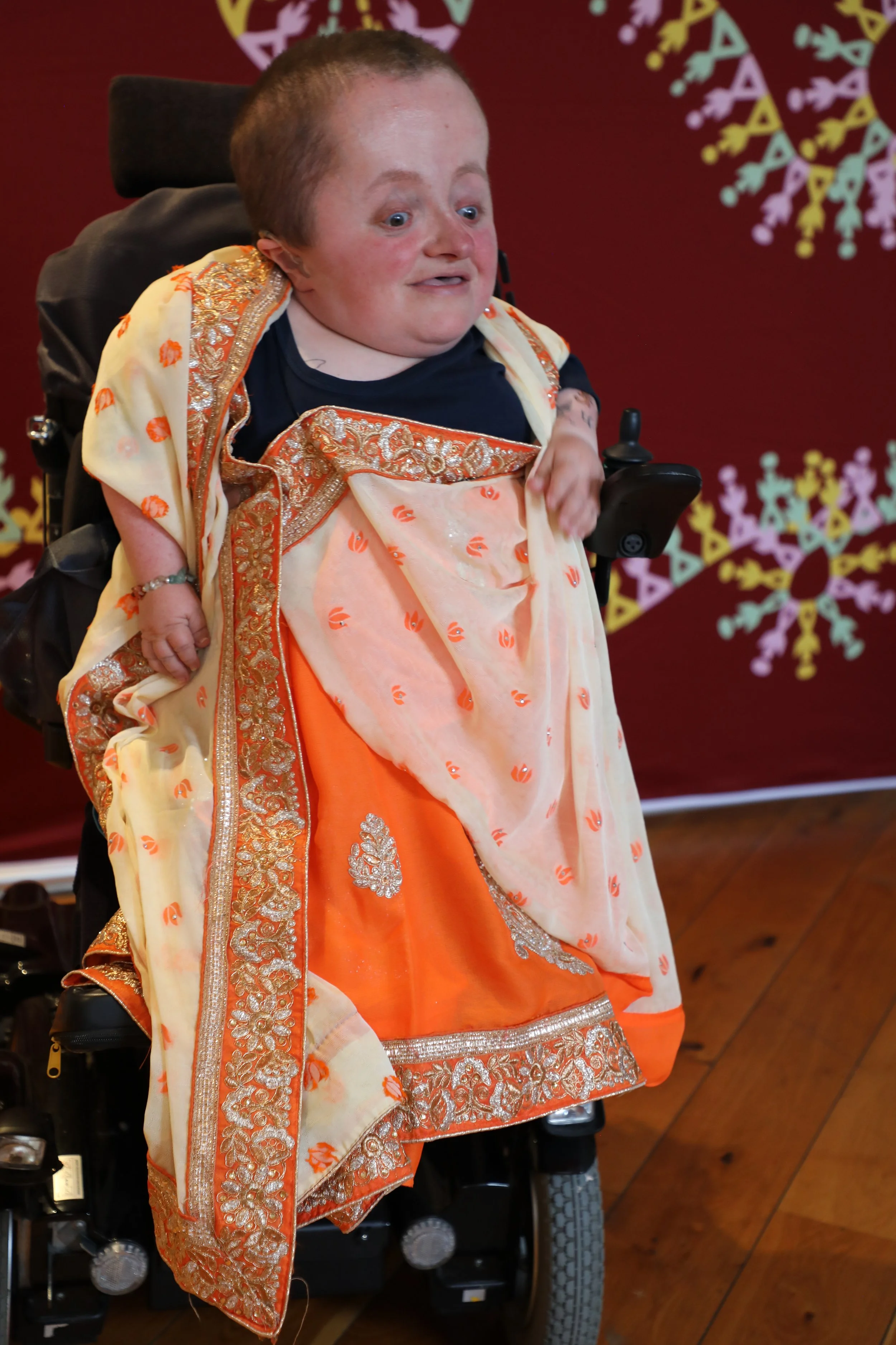 A person dressed in an orange patterned saree sitting in a power chair 