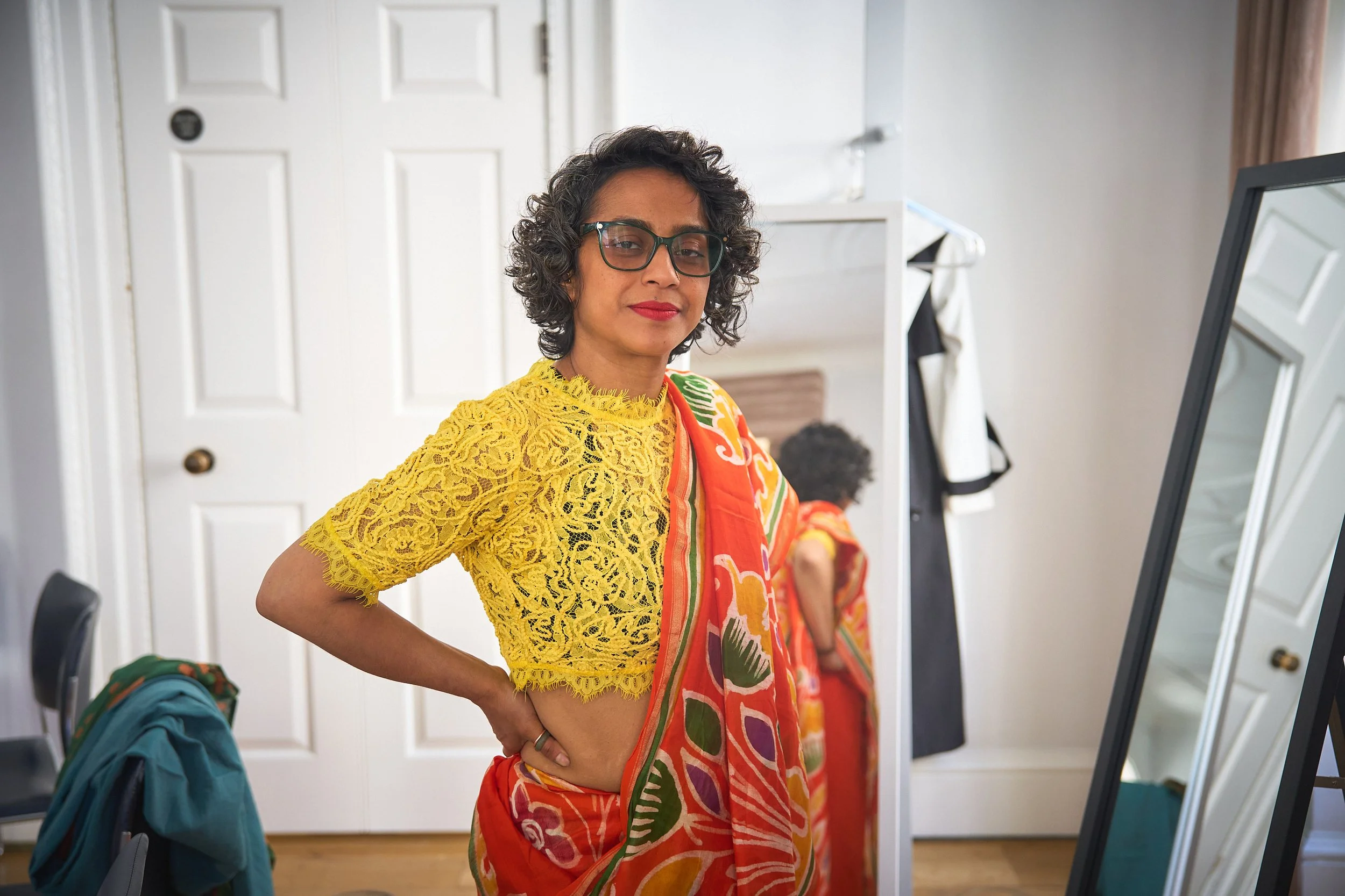 Poppy, a British-Bangladeshi woman stands with one hand on her hip, wearing an orange saree and yellow blouse