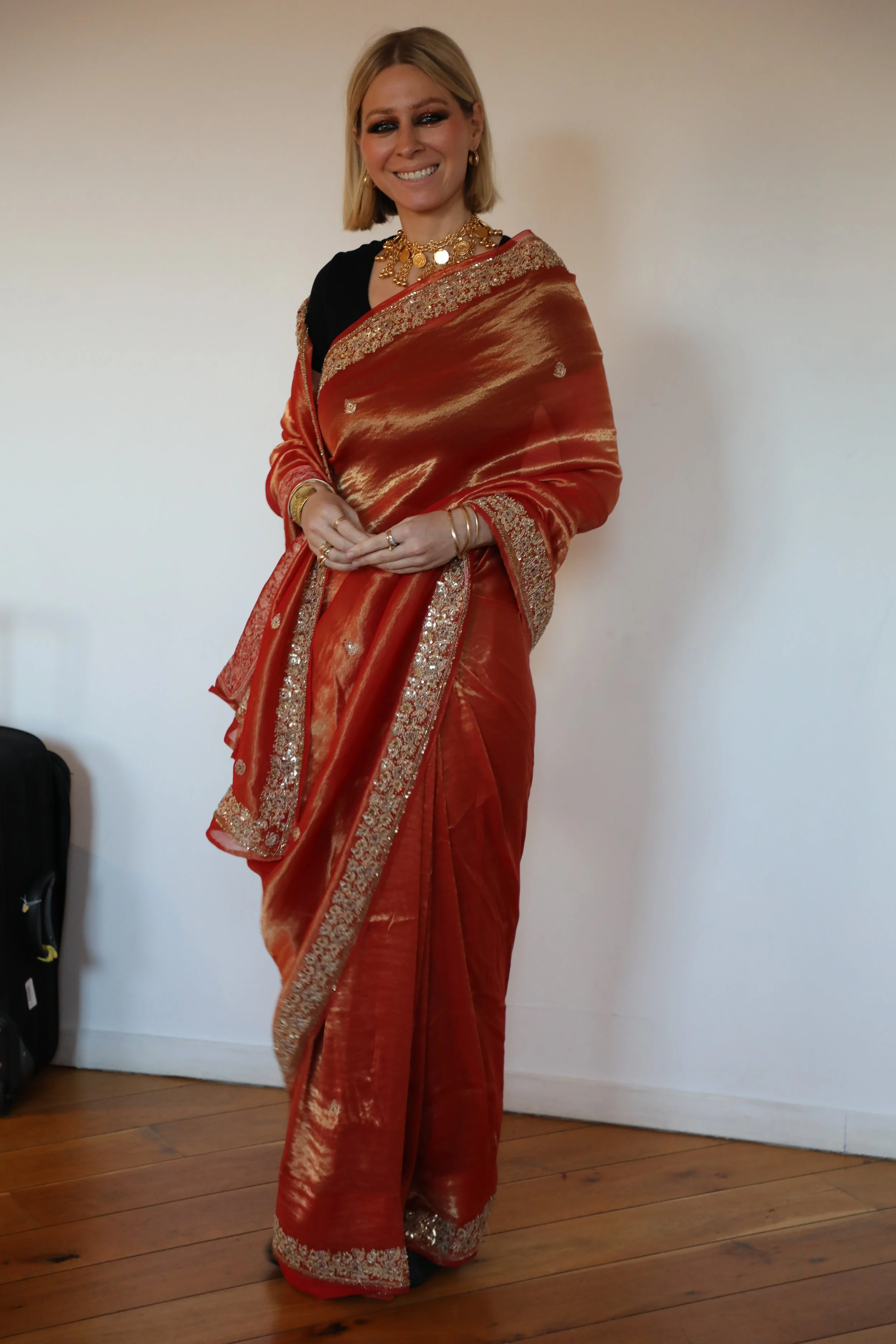 A woman wearing an orange saree stands and smiles at the camera