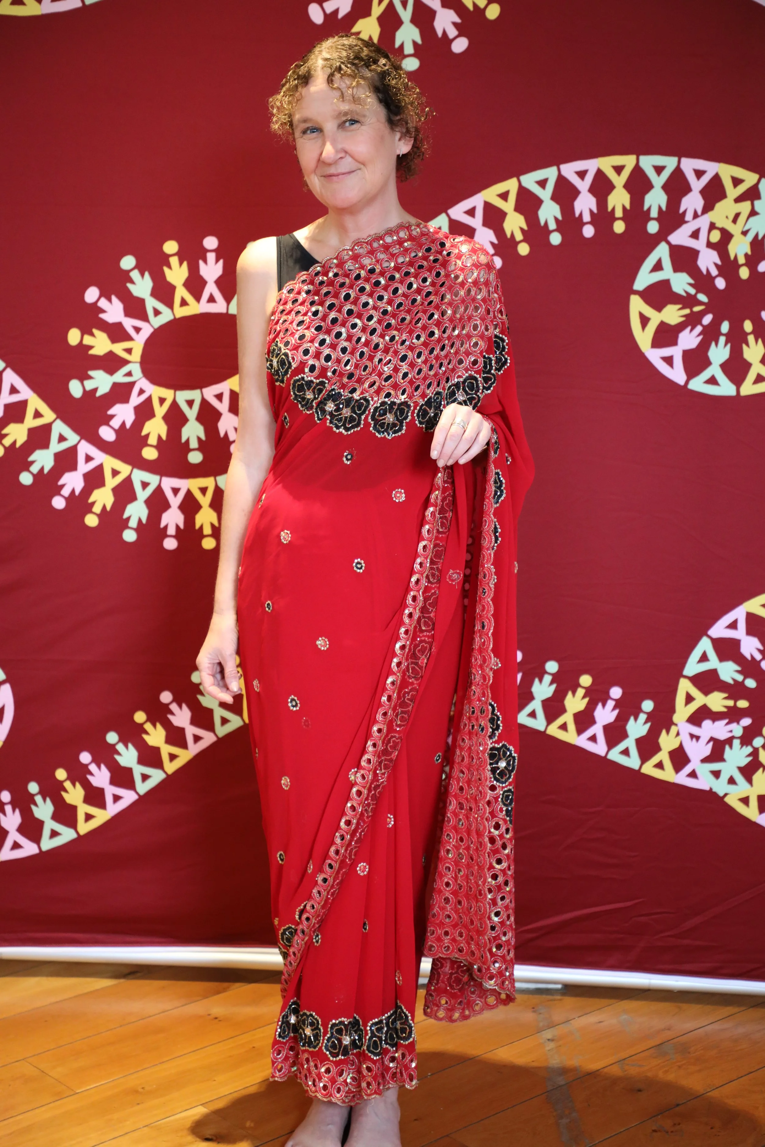 A woman with curly brown hair dressed in a red and green saree smiles at the camera
