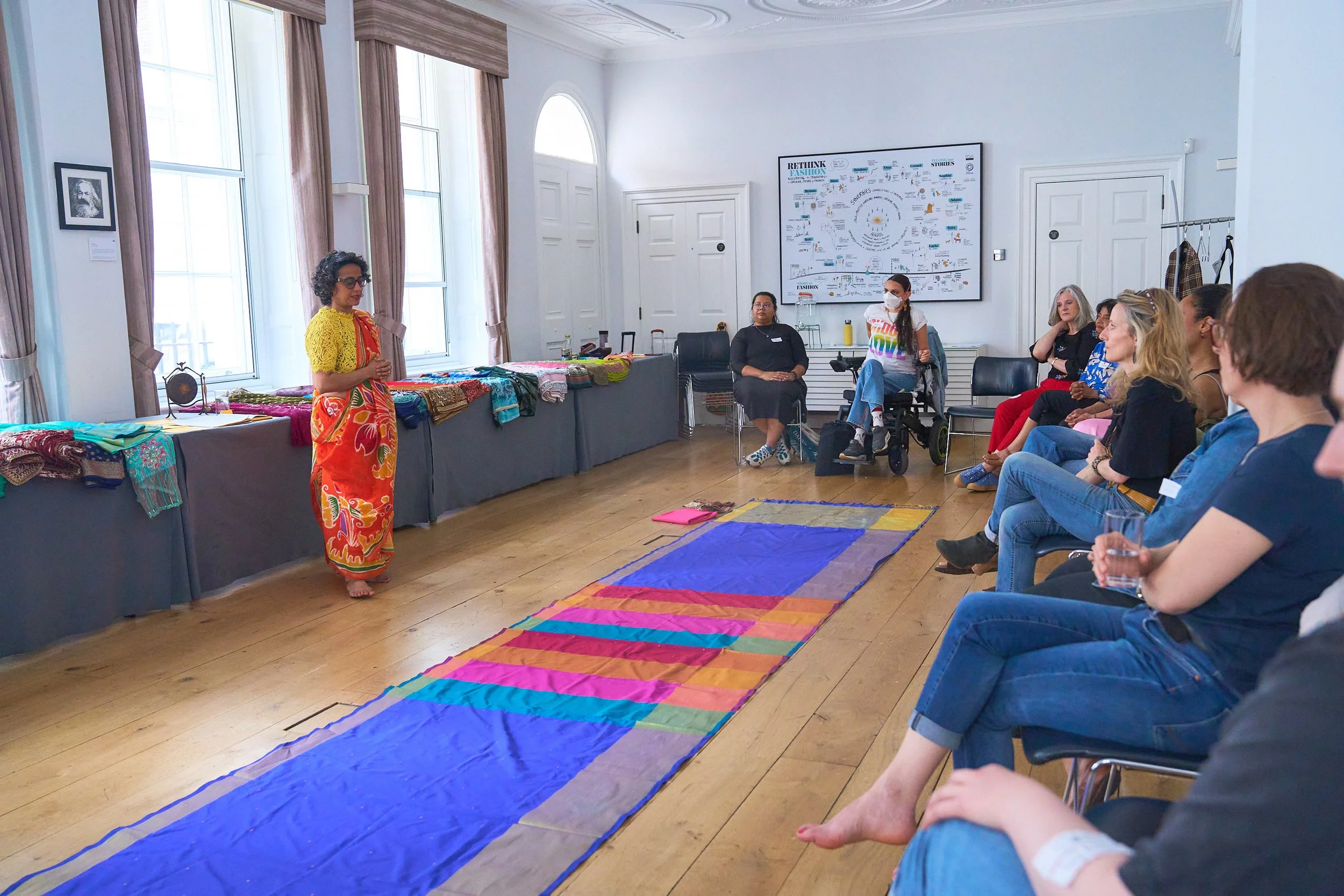 a group of women sit in a semi-circle, in front of them is a blue patterned saree laid out on the floor. Poppy stands at the front of the room