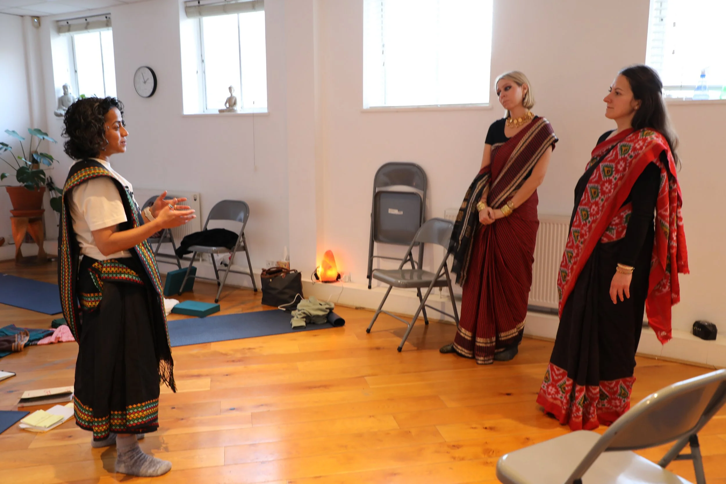 A woman with dark curly hair wearing a black saree stands in front of two women wearing sarees and speaks to them