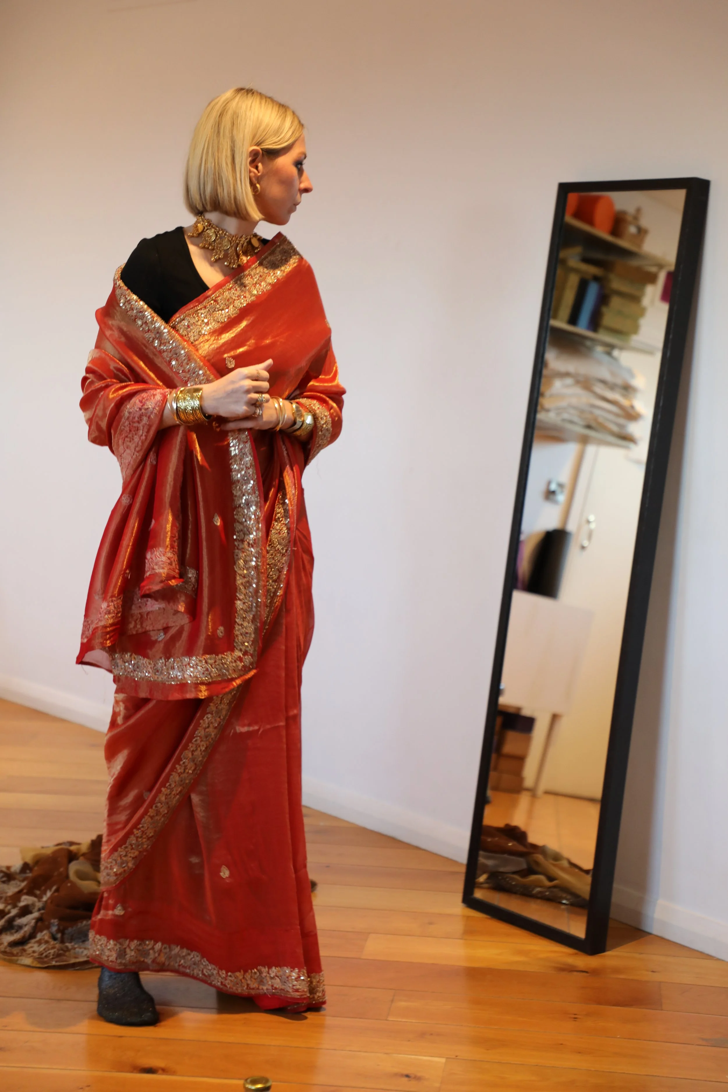 A blonde woman wearing an orange saree looks at her reflection in the mirror