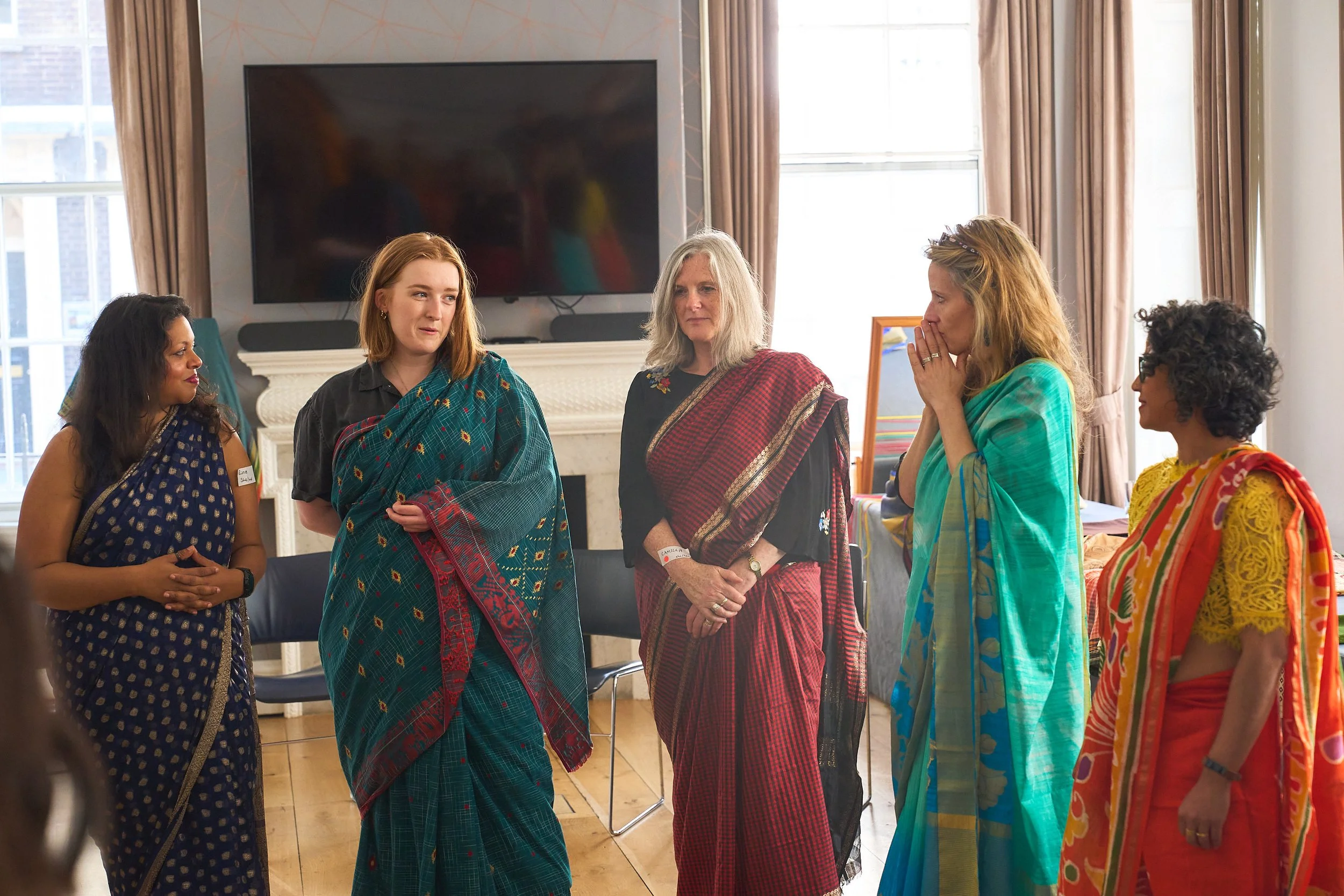 five women in sarees, of different ethnicities, stand together and talk