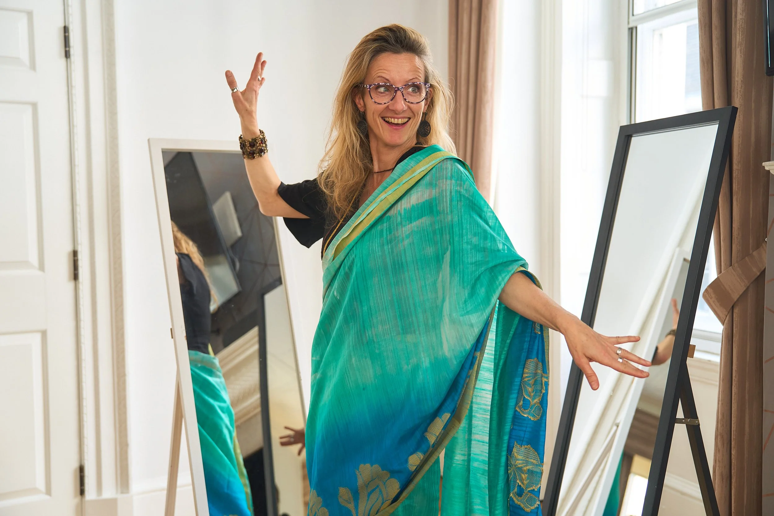 A white British woman wearing a teal saree poses with one hand raised and the other flared out.