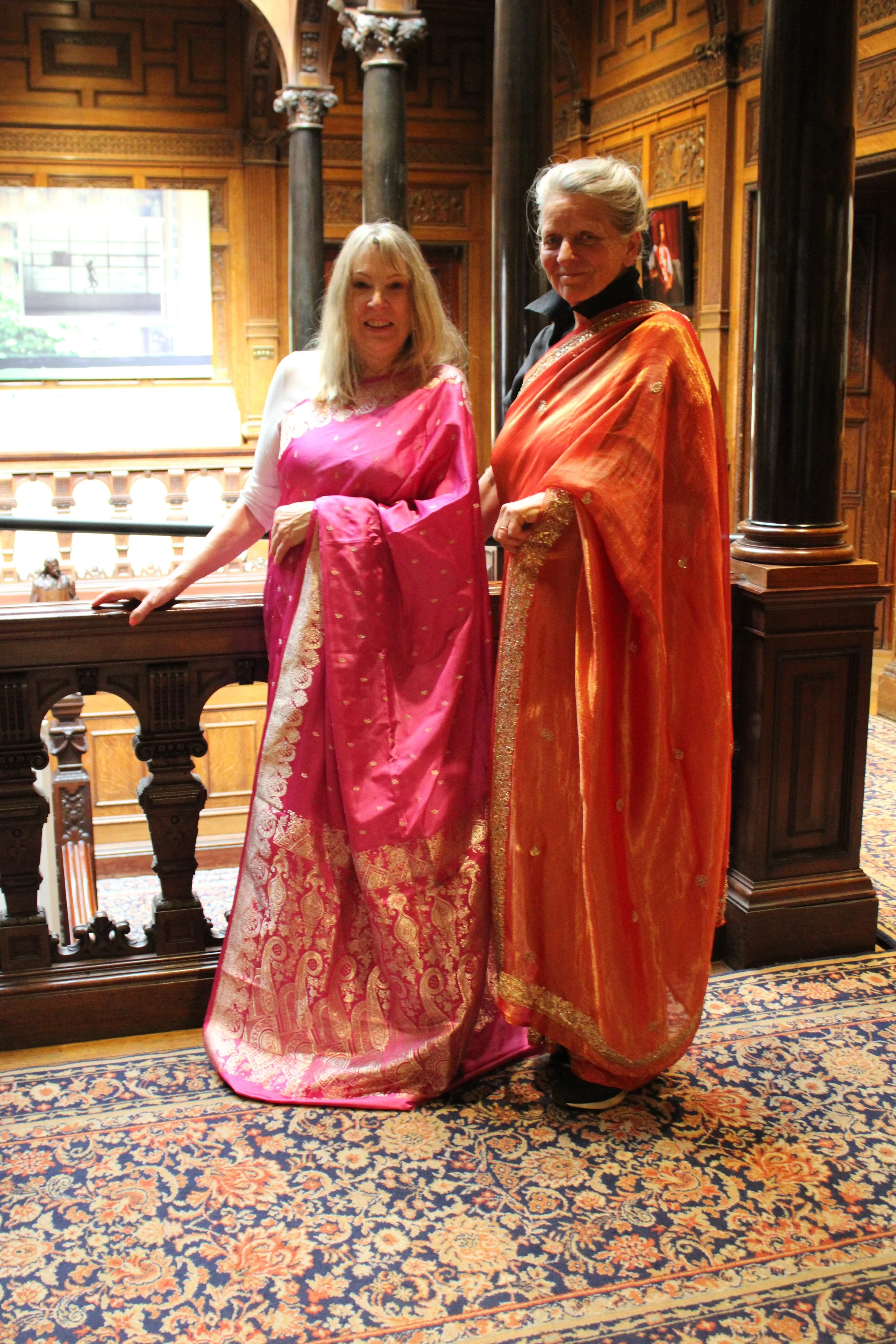 Two white British women wearing pink and orange sarees smile at the camera
