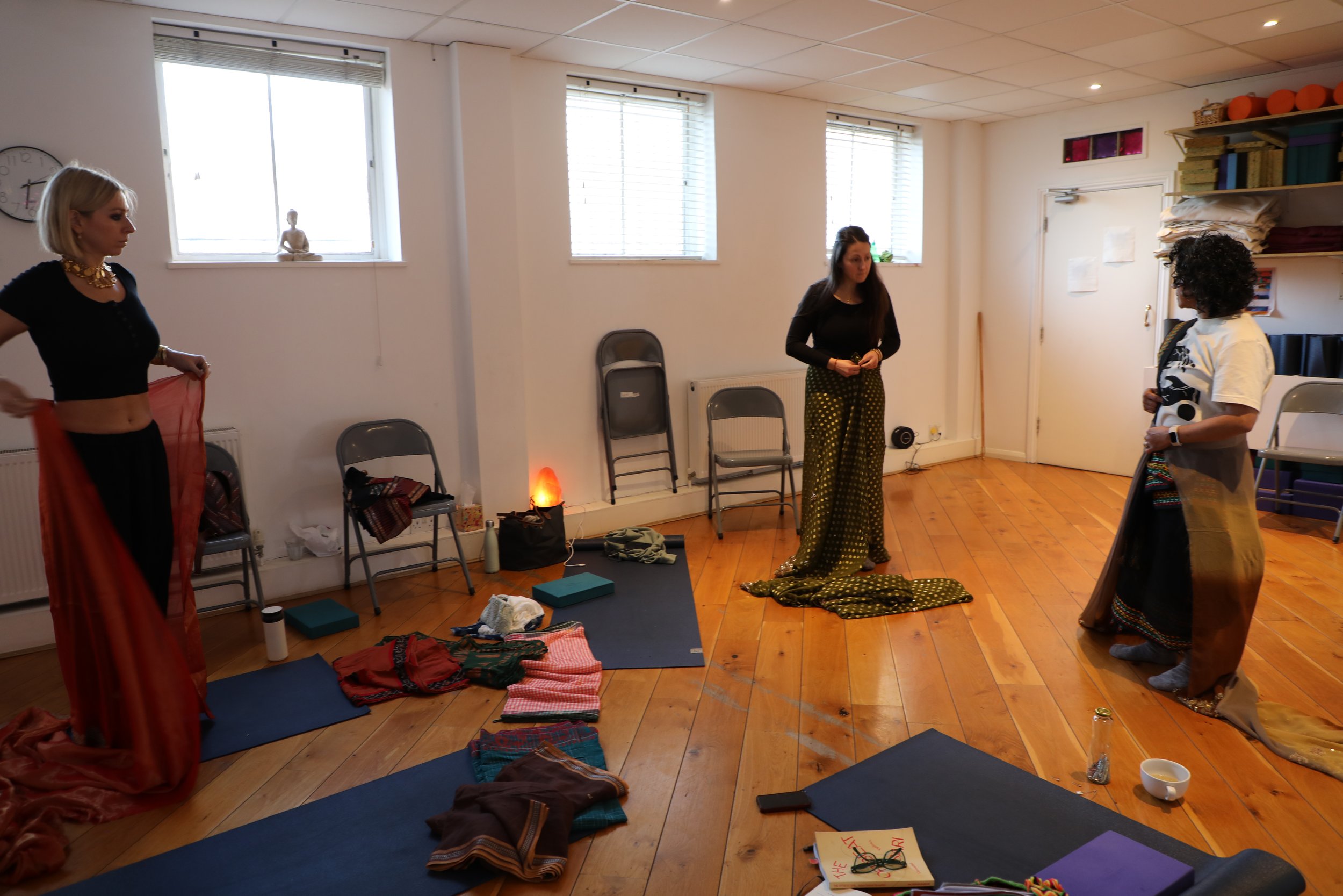 Three women drape sarees on themselves in a room with yoga mats and other sarees on the floor