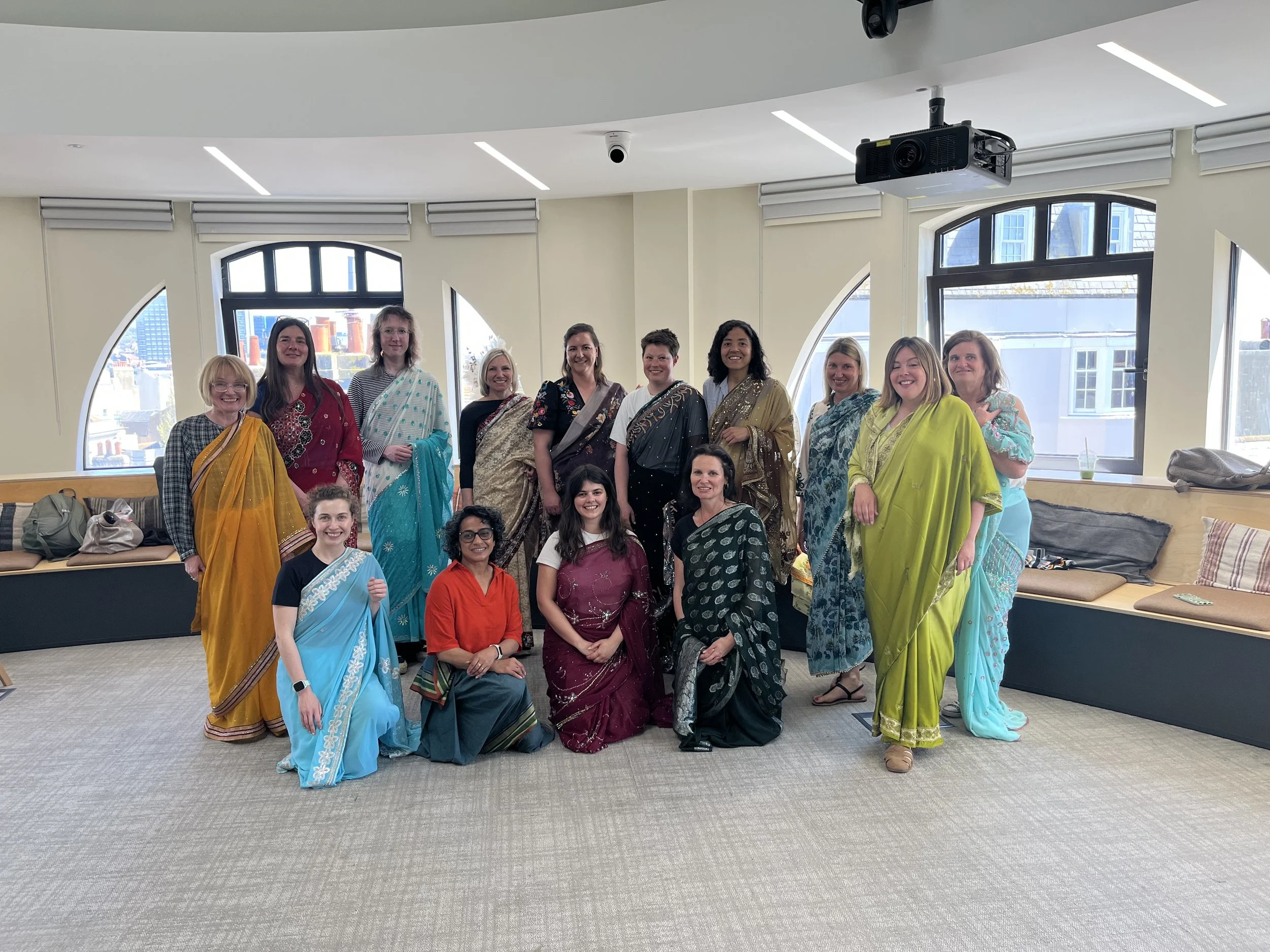 A group of women wearing colourful sarees smile at the camera, some standing up with four women kneeling in front of them