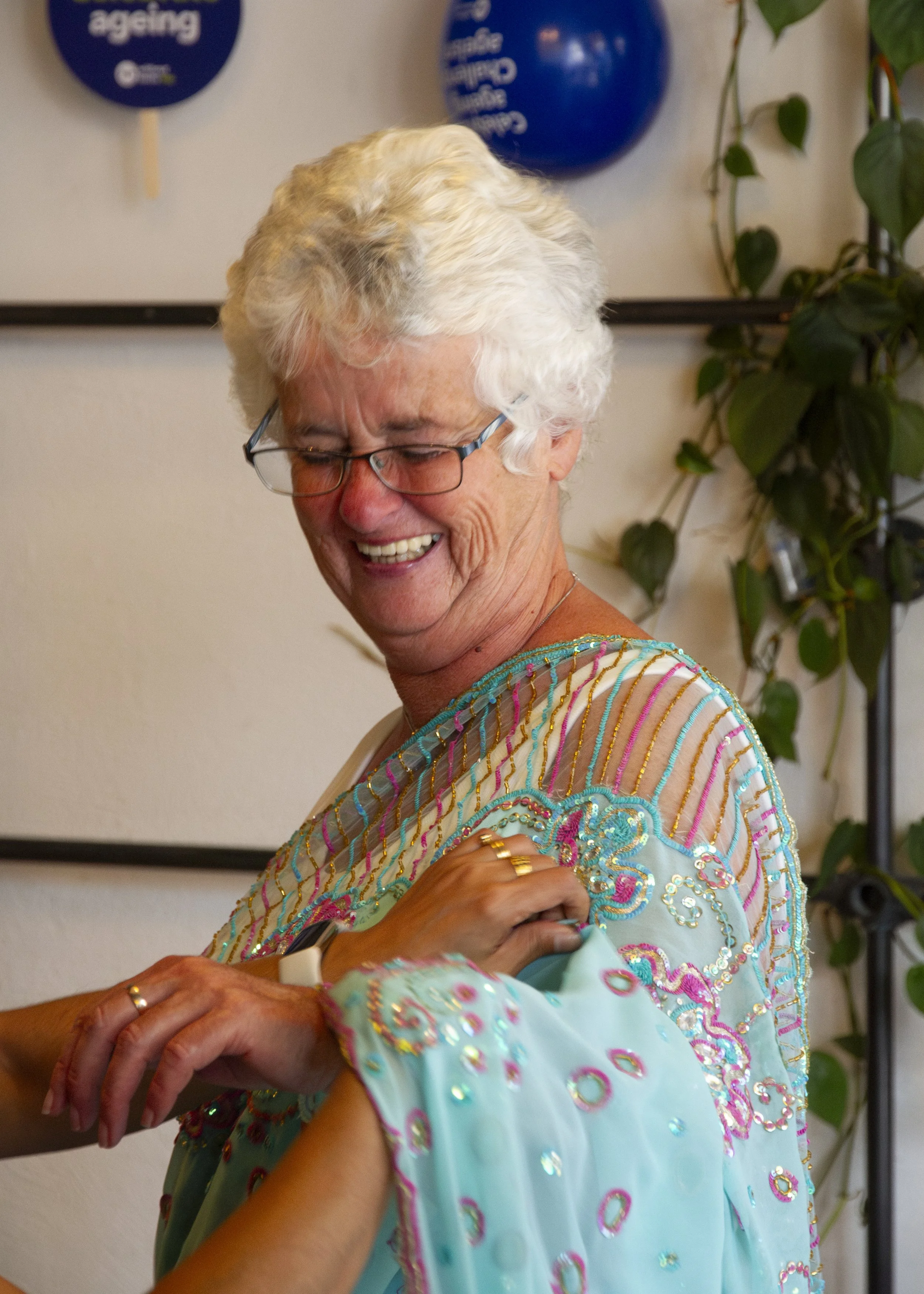 A White British woman with curly white hair smiles and laughs while being helped to dress in a saree