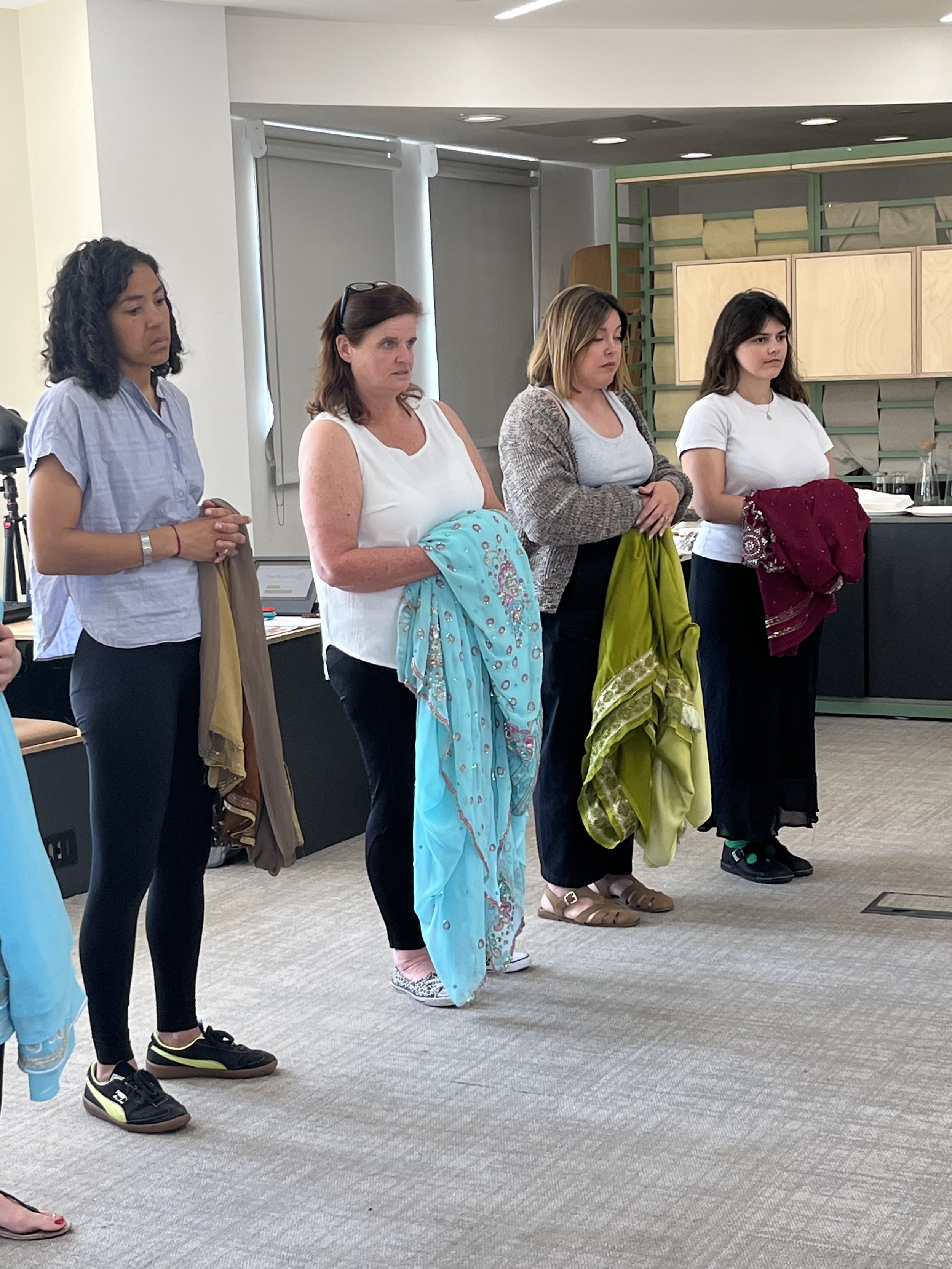 Four women holding sarees look toward the front of the room, listenting to a speaker