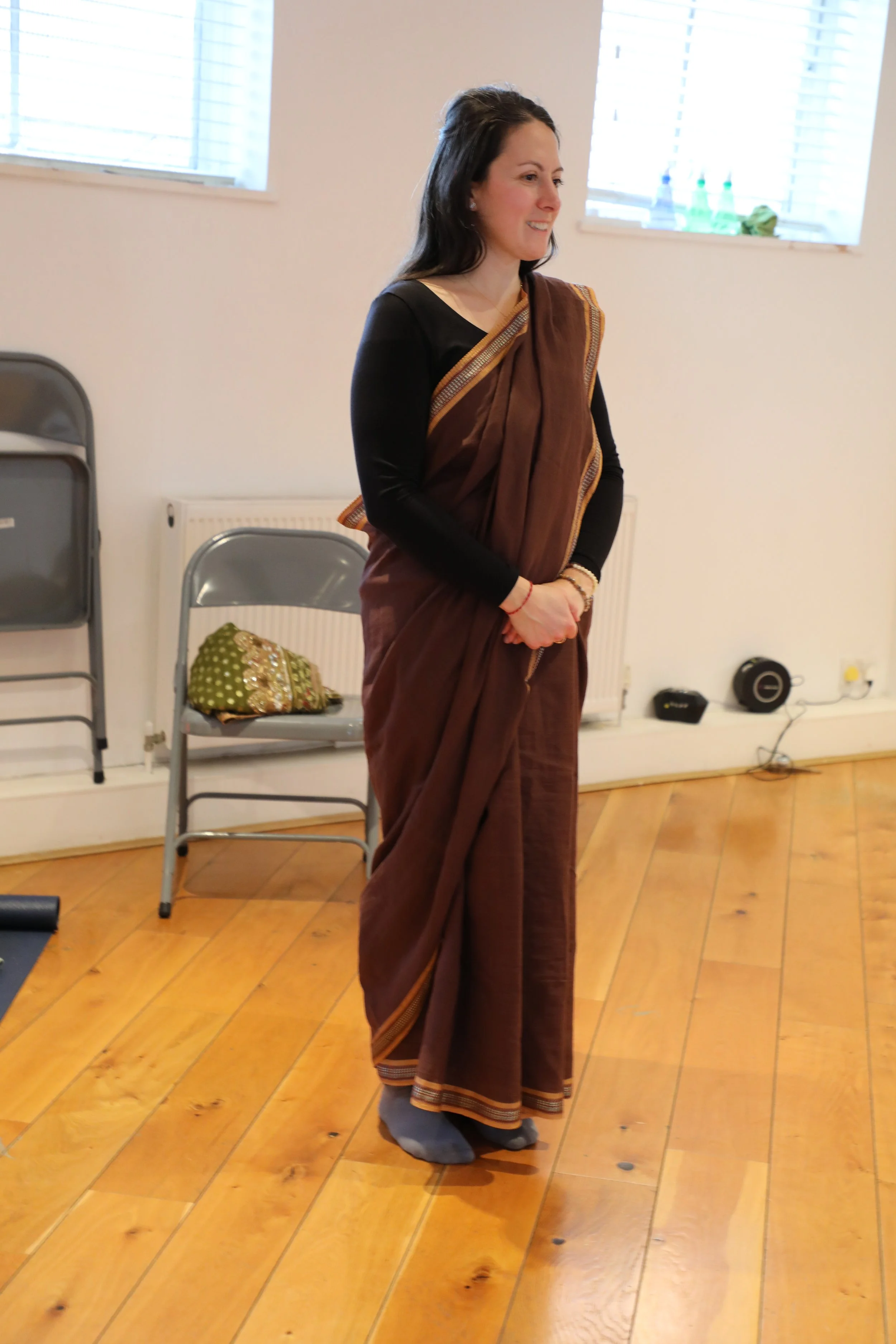 A woman wearing a black top and brown saree stands with her hands clasped and smiles