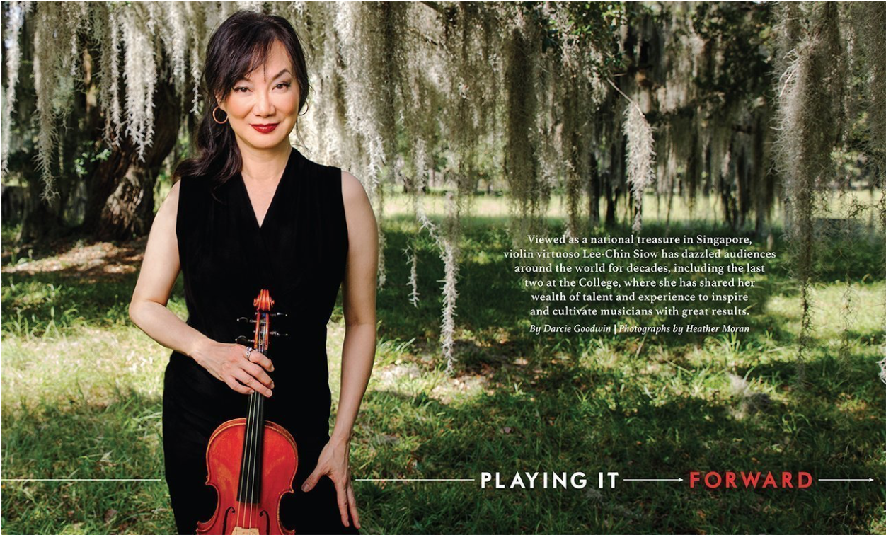 A woman holding a violin standing outdoors near a tree with hanging moss, wearing a black sleeveless dress and smiling.