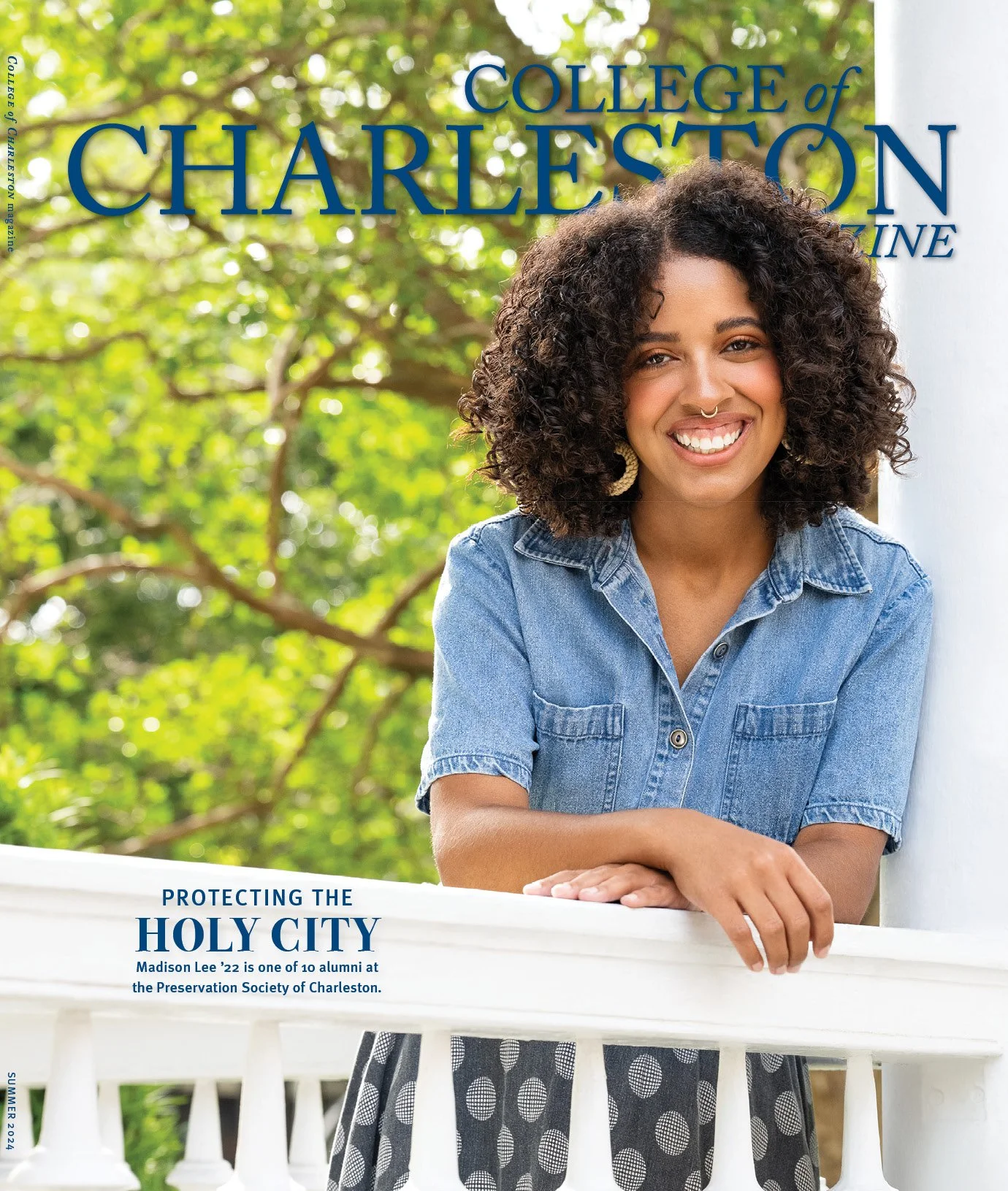 A young woman with curly hair smiling, wearing a denim shirt, standing outdoors with green trees in the background and a white railing in front of her, related to College of Charleston.