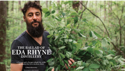 A man with a beard standing behind green foliage in a forest setting, holding onto a bush, with a book titled 'The Ballad of Eda Rhyne Distillery' in front of him.