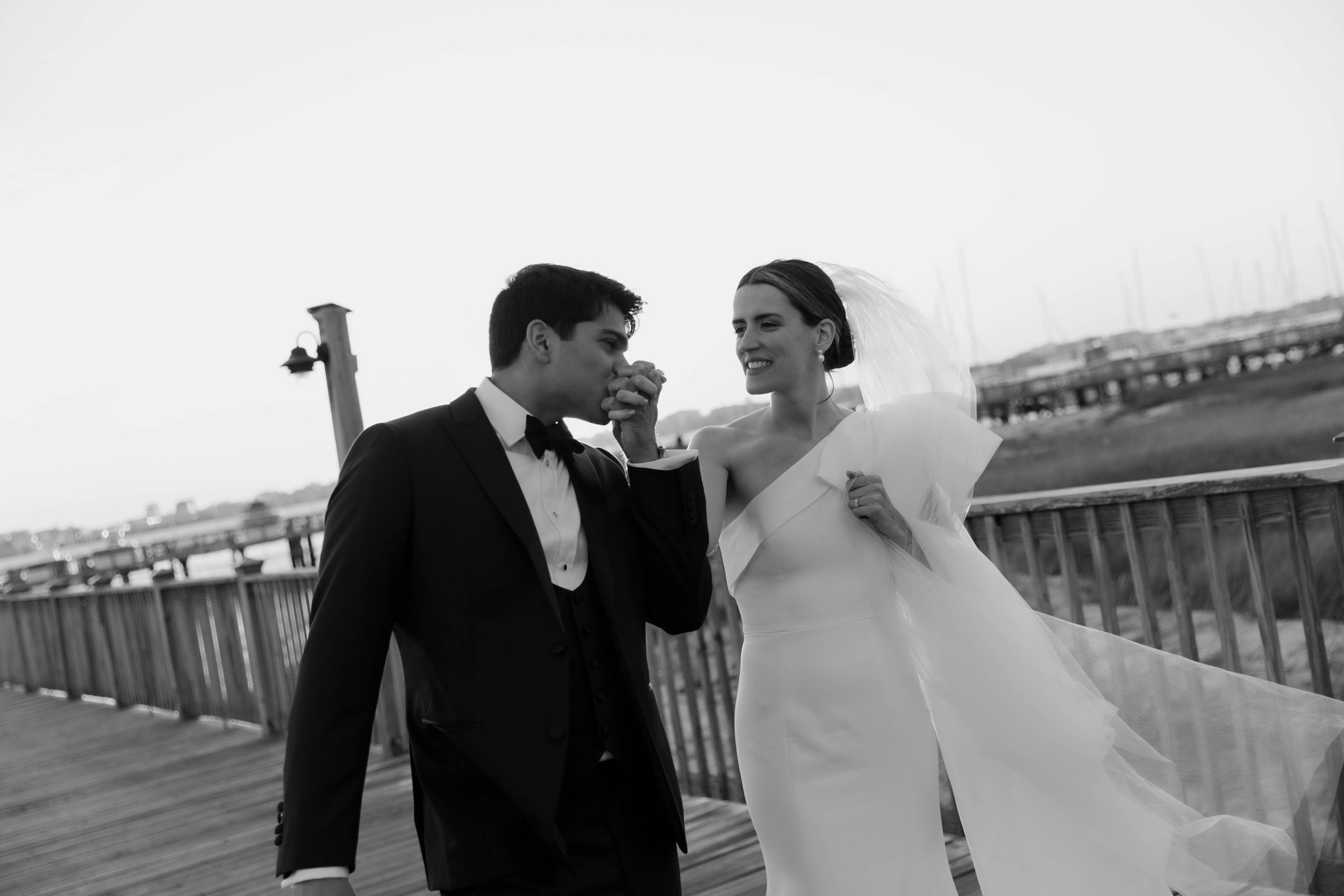 A black and white photo of a bride and groom on a pier, with the groom kissing the bride's hand while she smiles.