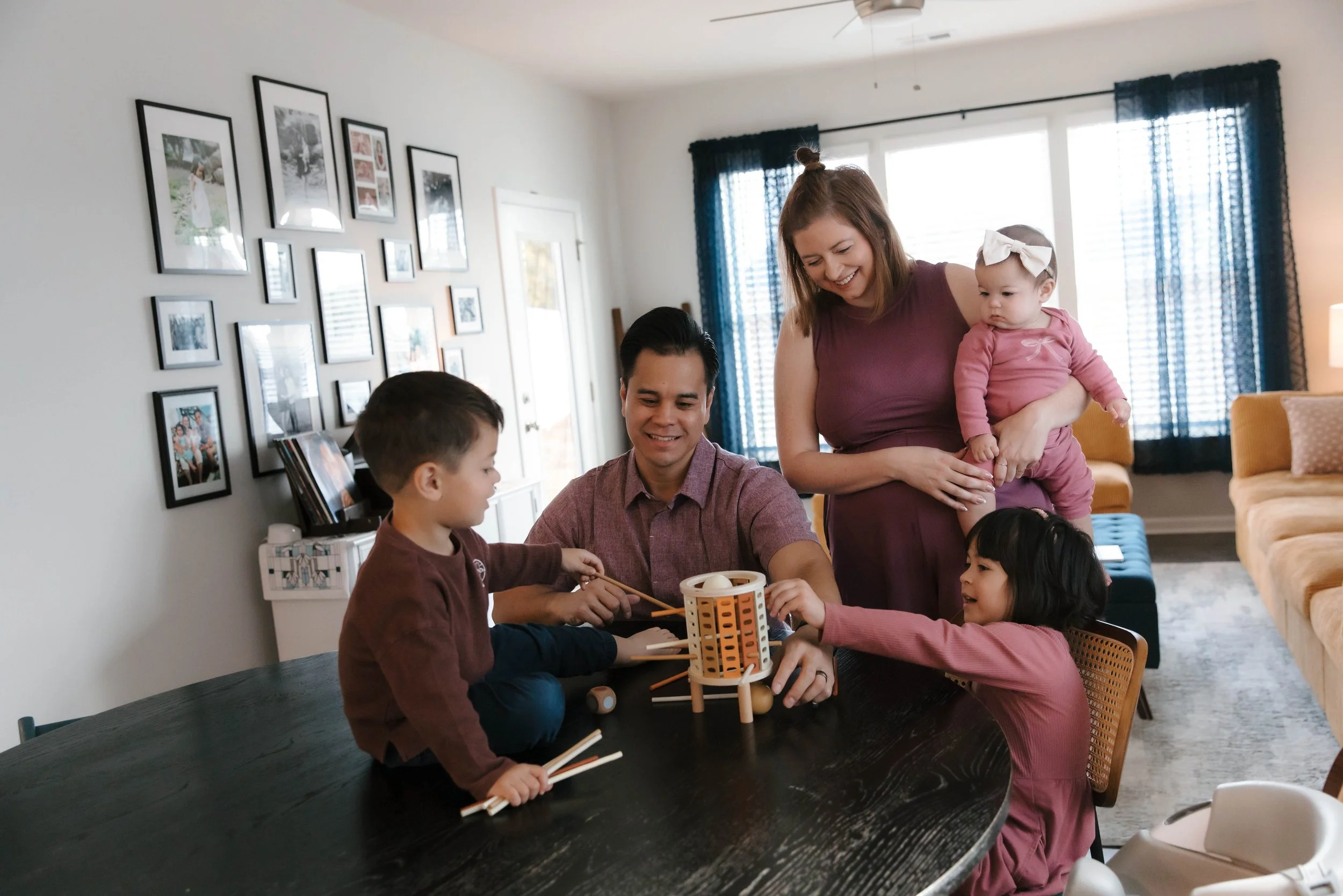 A family gathers around a dining table playing a game, with a woman holding a baby and smiling, in a bright living room with framed photos on the wall and large windows with blue curtains.