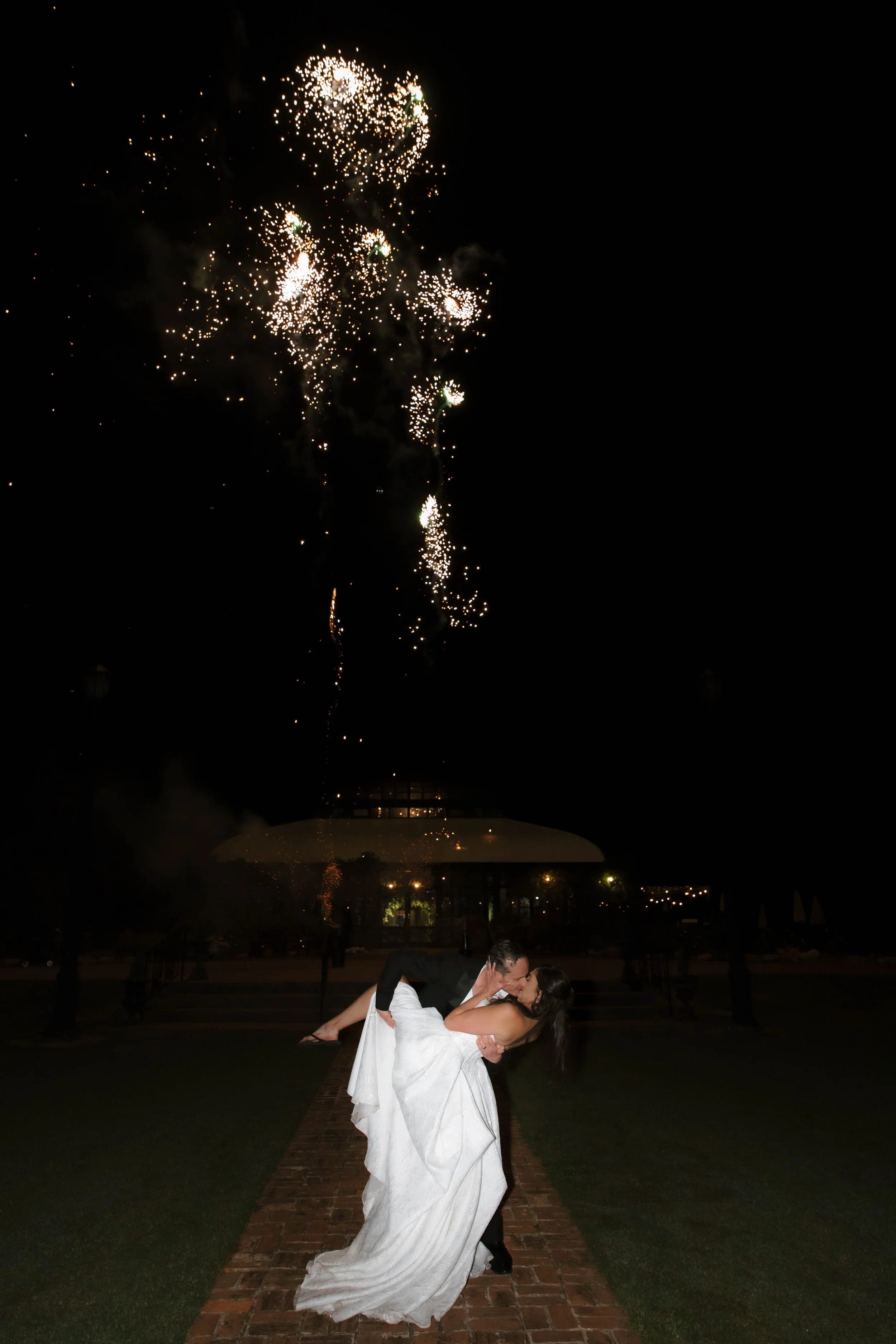 A bride and groom are dancing outdoors at night under fireworks in the sky.