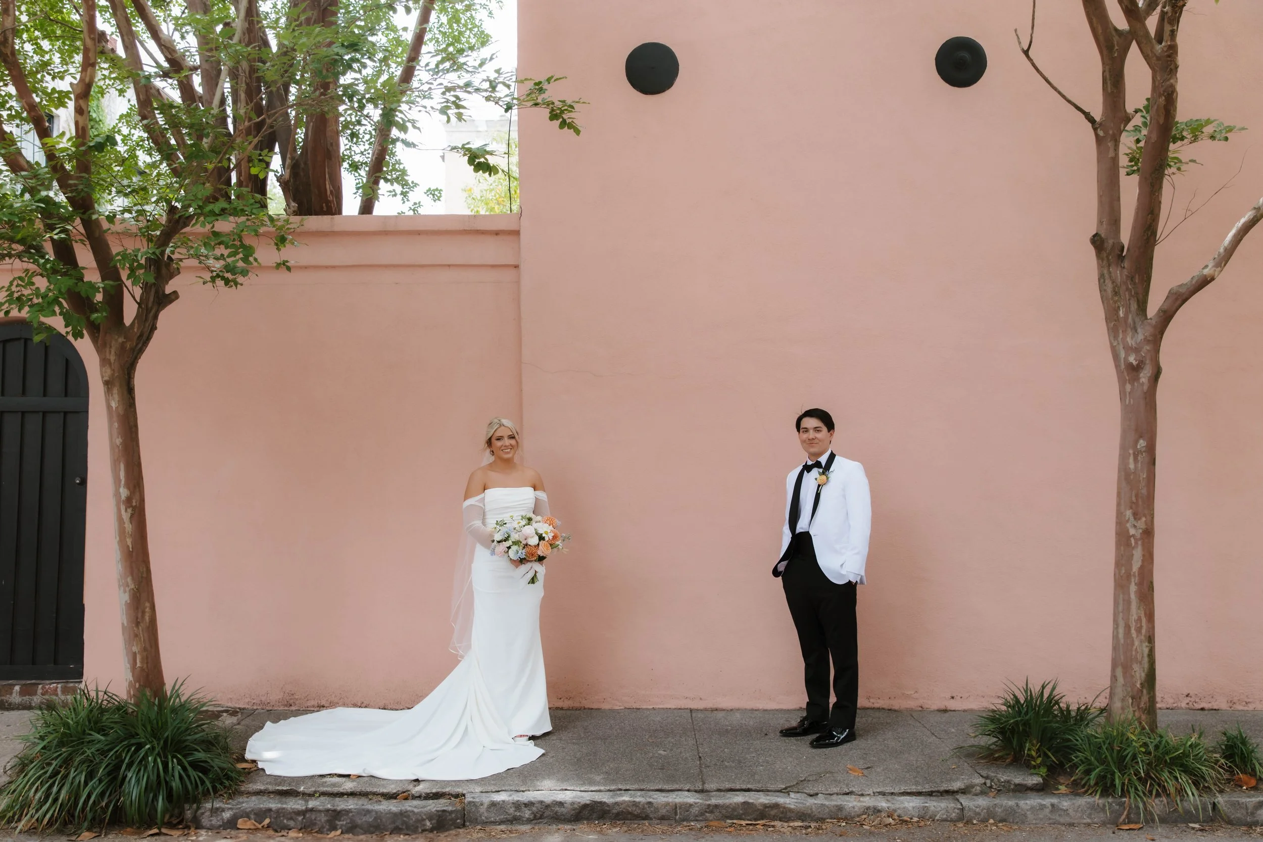 A bride and groom standing outdoors in front of a pink wall with two trees on either side. The bride is holding a bouquet of flowers, wearing a white wedding dress, and has blonde hair. The groom is dressed in a black and white tuxedo, with a white s