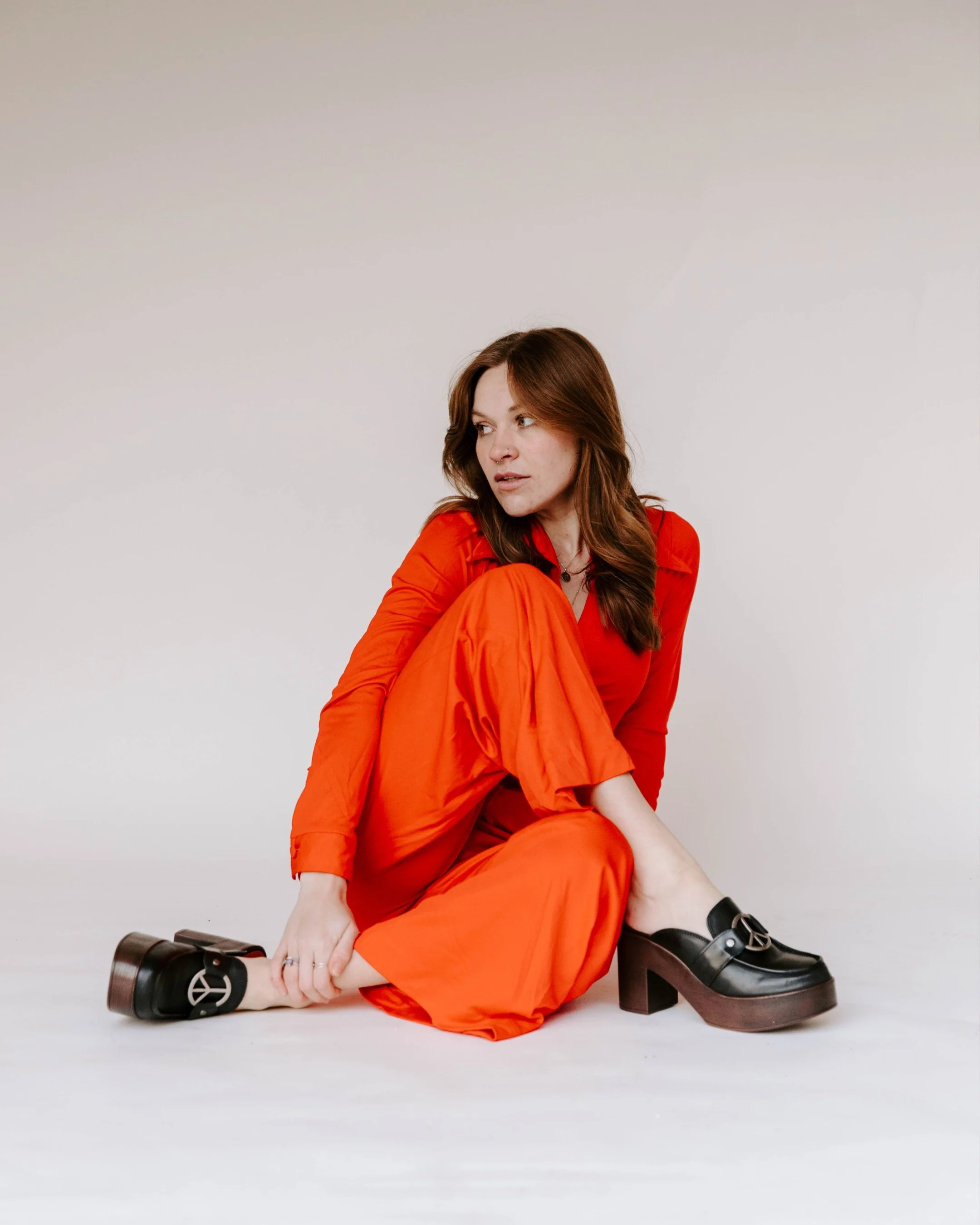 Photographer in a red outfit sitting on the floor, black platform shoes with buckle, against a plain light-colored background.