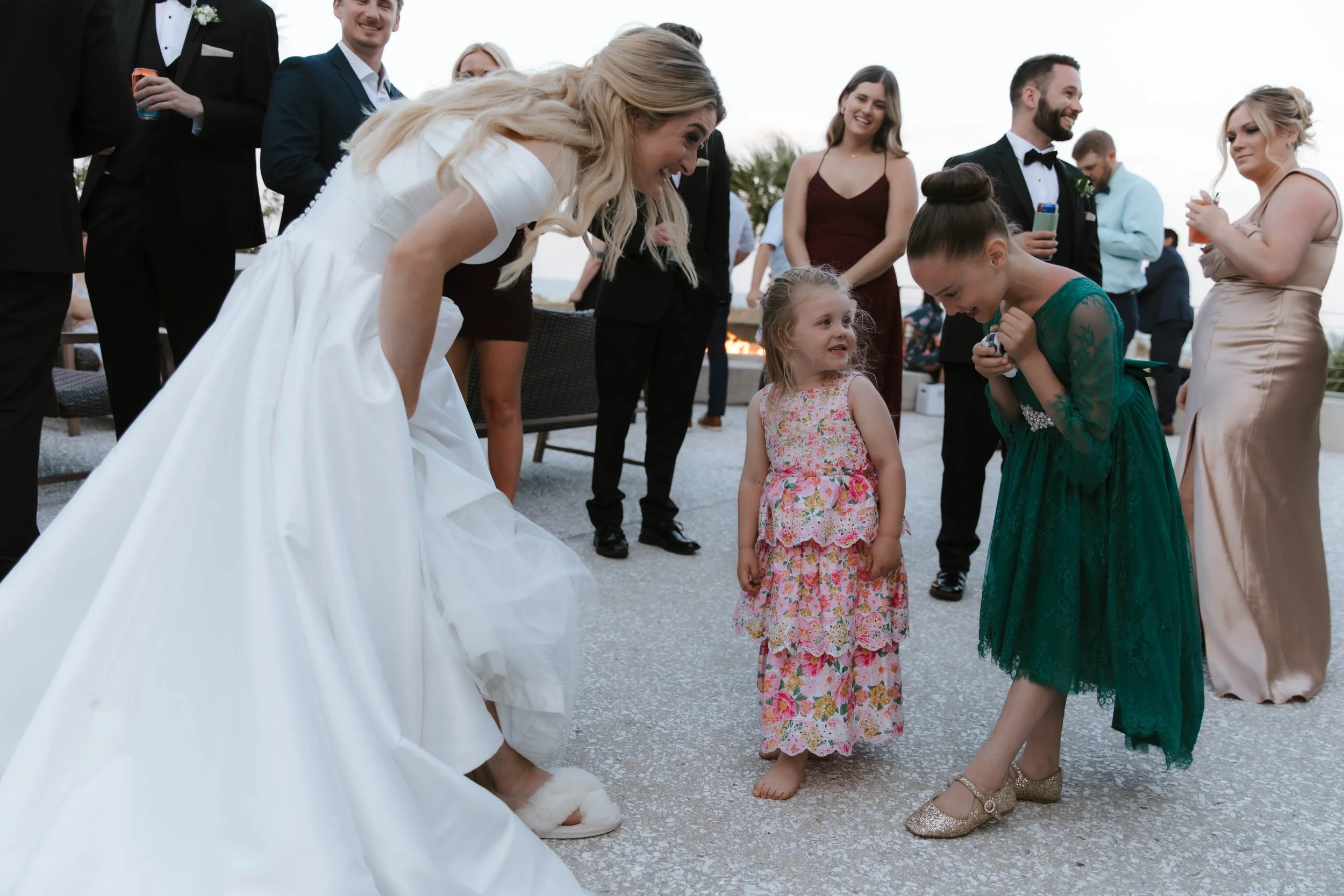 Group of people in formal attire gathered outdoors at a wedding celebration, with a woman in a white dress bending down to interact with a young girl in a floral dress, while others observe and smile.